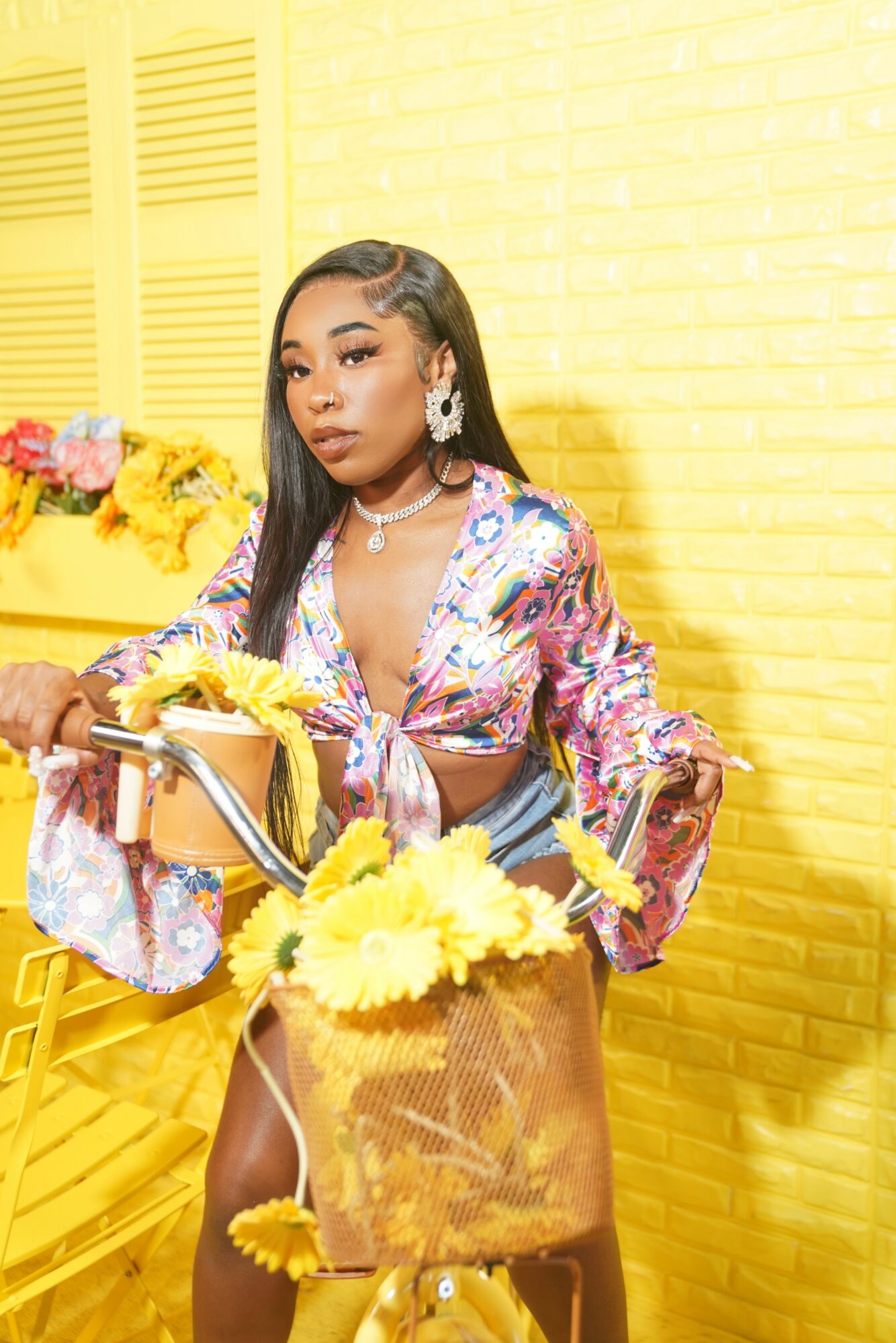 Woman with long hair and earrings stands in front of a yellow wall with flowers, holding a bicycle with a yellow bag.