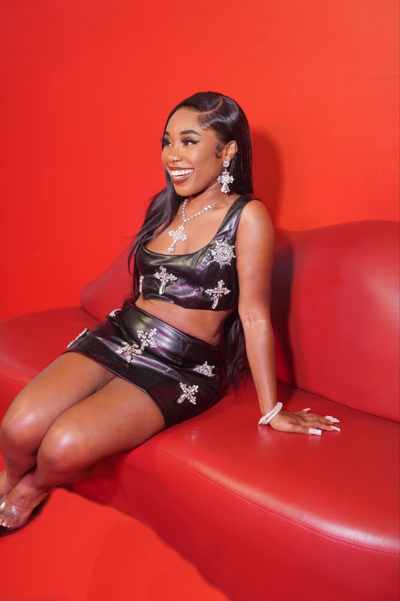 Woman sitting on red couch, smiling, wearing a black dress with silver embroidery, jewelry, and earrings.