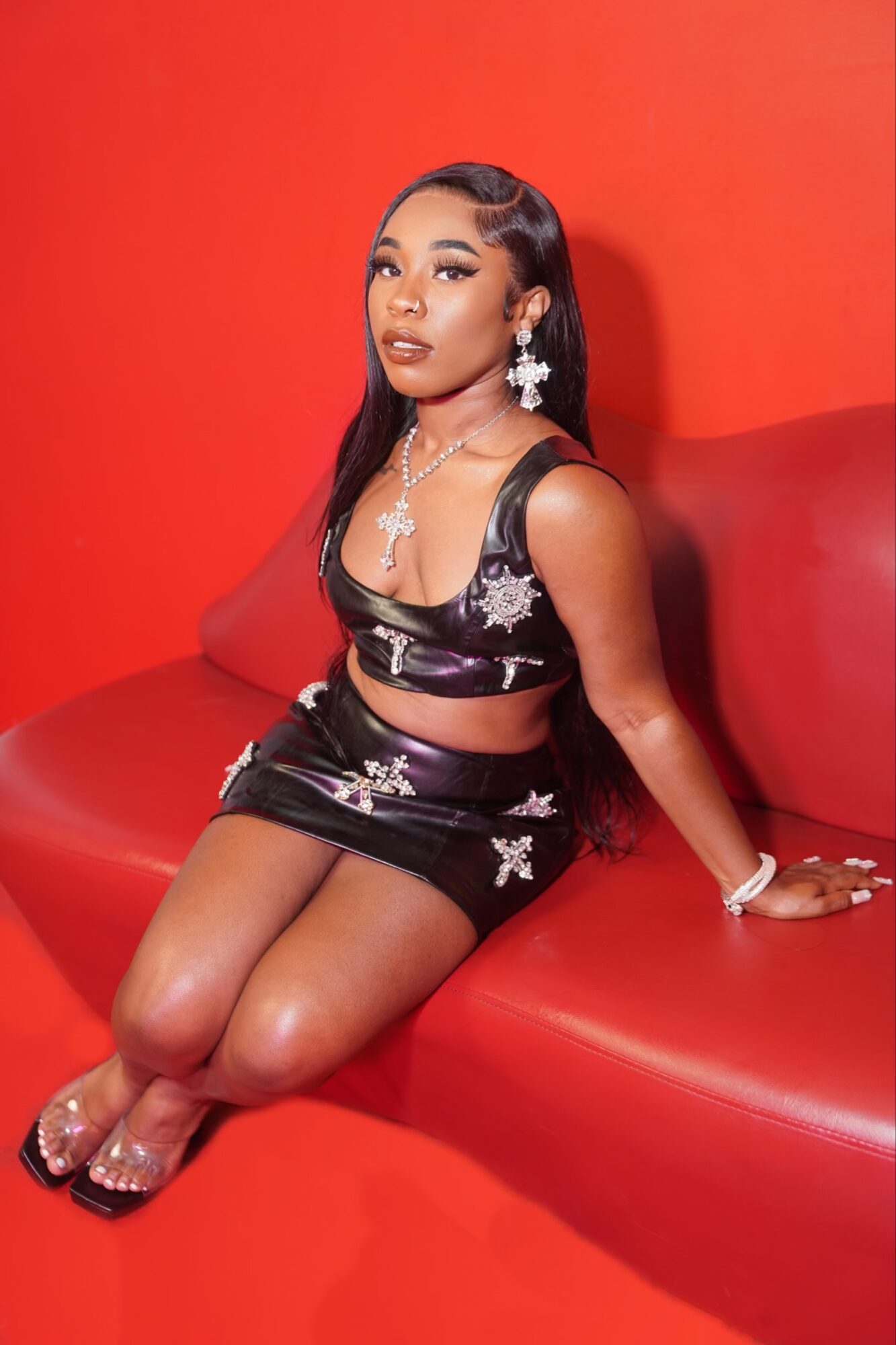 Young woman in black floral outfit sitting on red bench against red background, wearing jewelry and makeup, looking at camera.