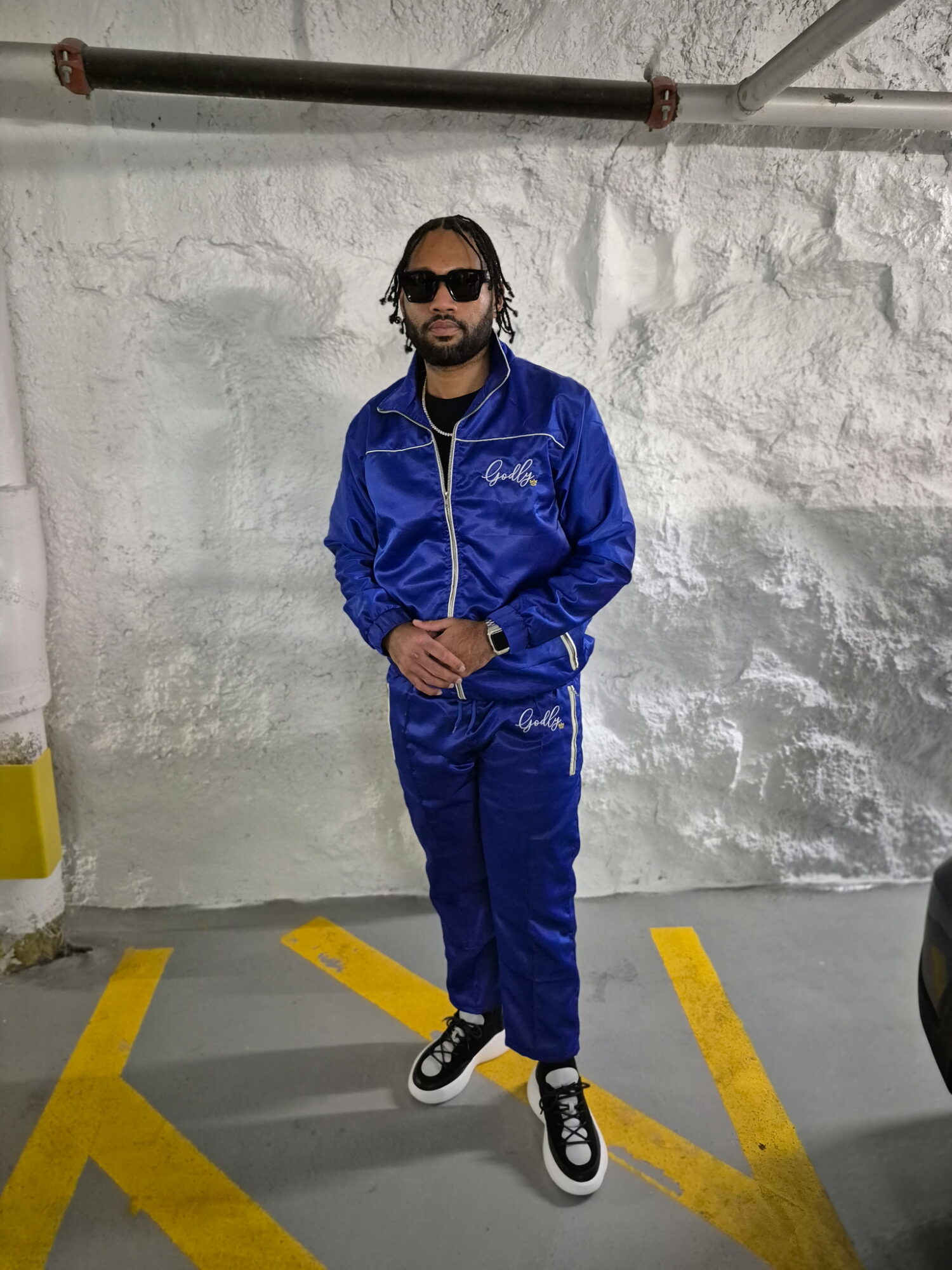 Man with sunglasses and braided hair in a blue tracksuit standing in a parking lot against a white wall.