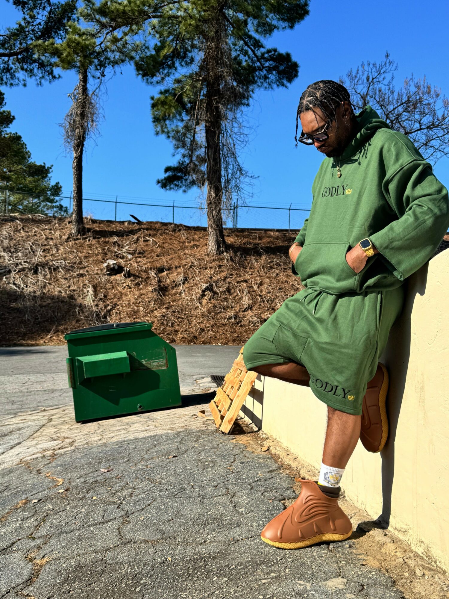 Person in green coat and boots standing outdoors near a green box, with trees and blue sky in background.