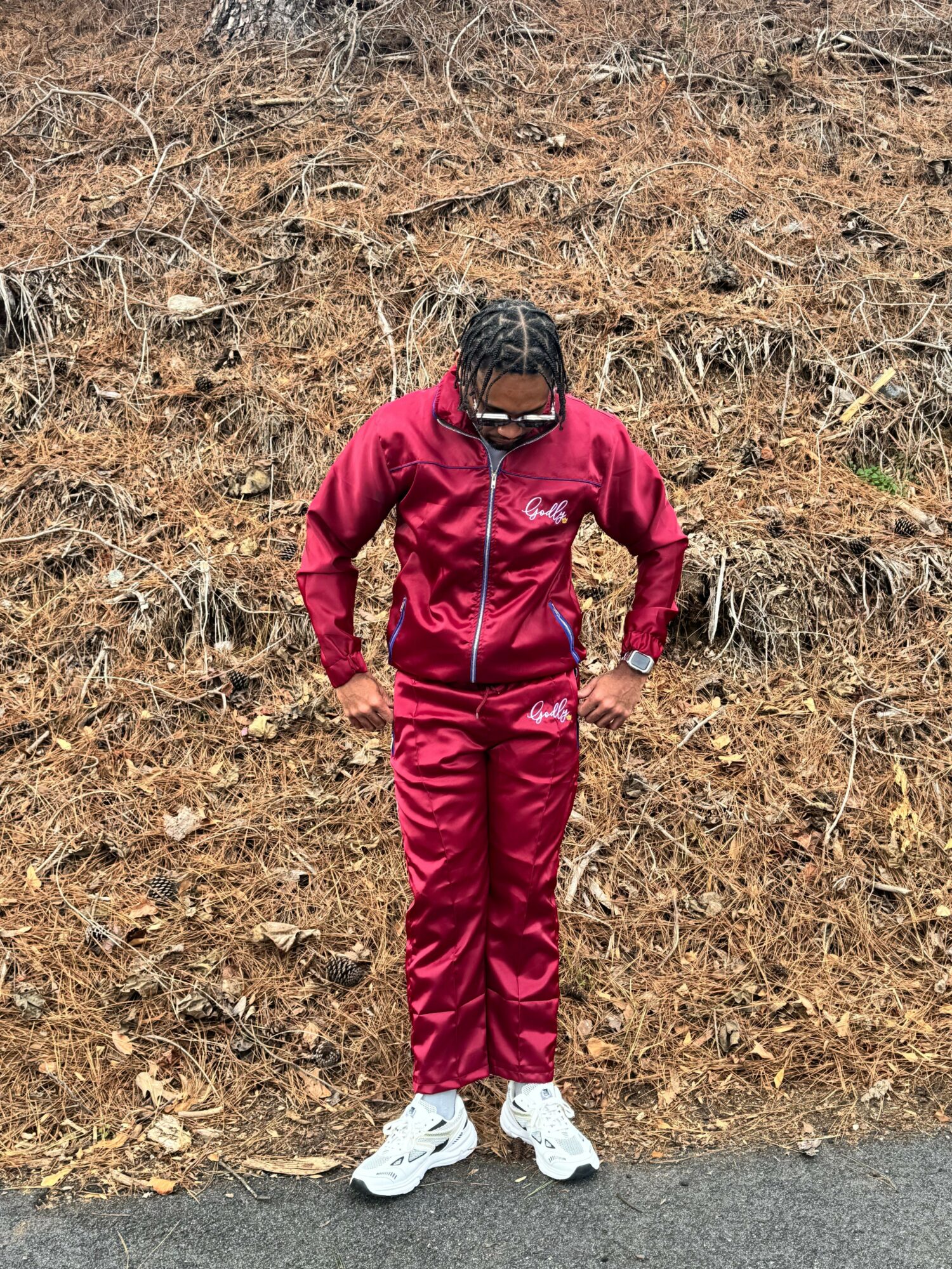Person in red tracksuit and white shoes standing outdoors on dry grass, wearing sunglasses and a black cap.