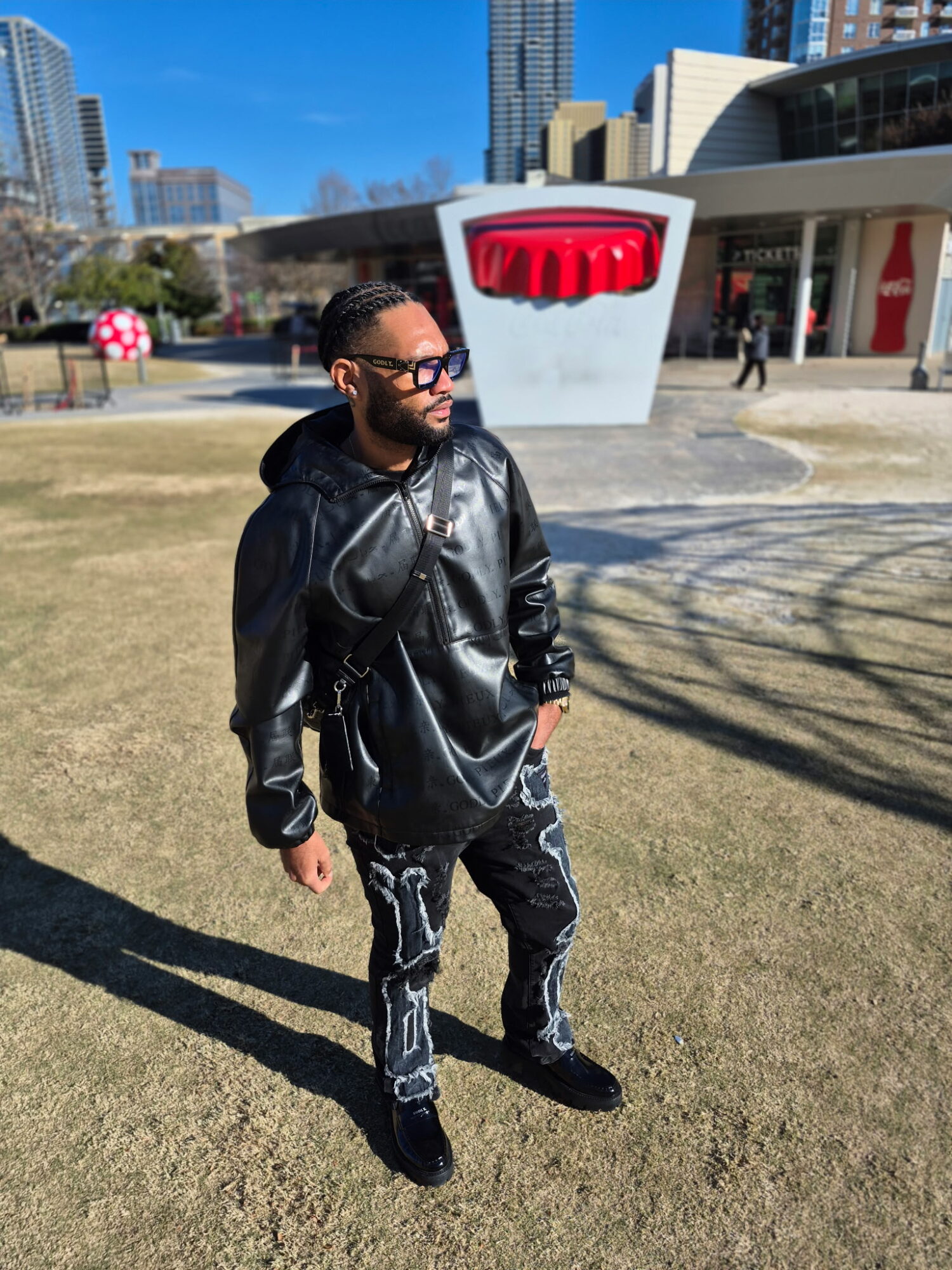 Man with sunglasses and a beard wearing a black leather jacket and patterned pants standing outdoors in a city park.
