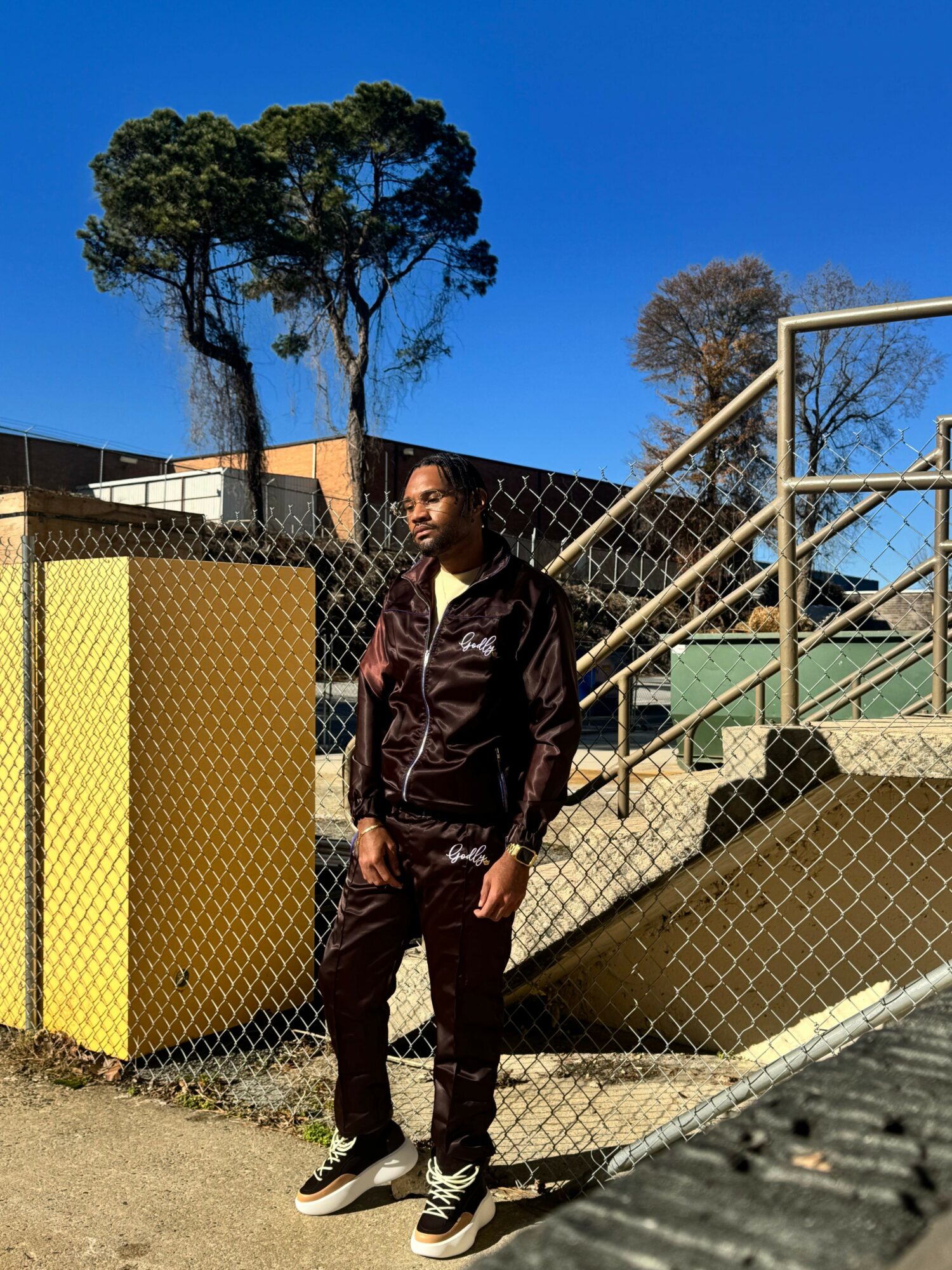 Person standing outdoors near a chain-link fence with trees and a blue sky in the background.