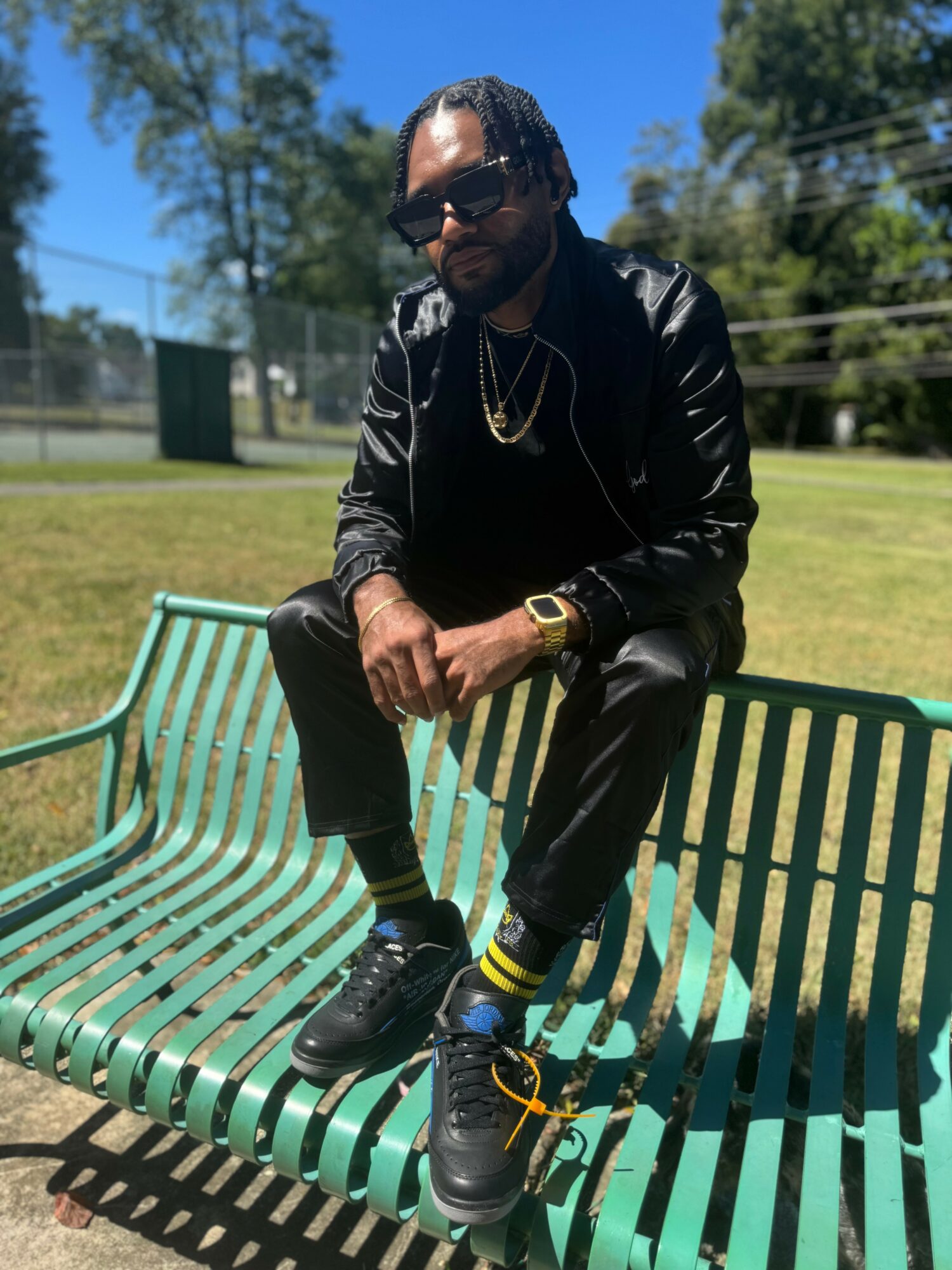 Person sitting on a green park bench outdoors, wearing sunglasses, black leather jacket, and sneakers, with trees and blue sky in background.