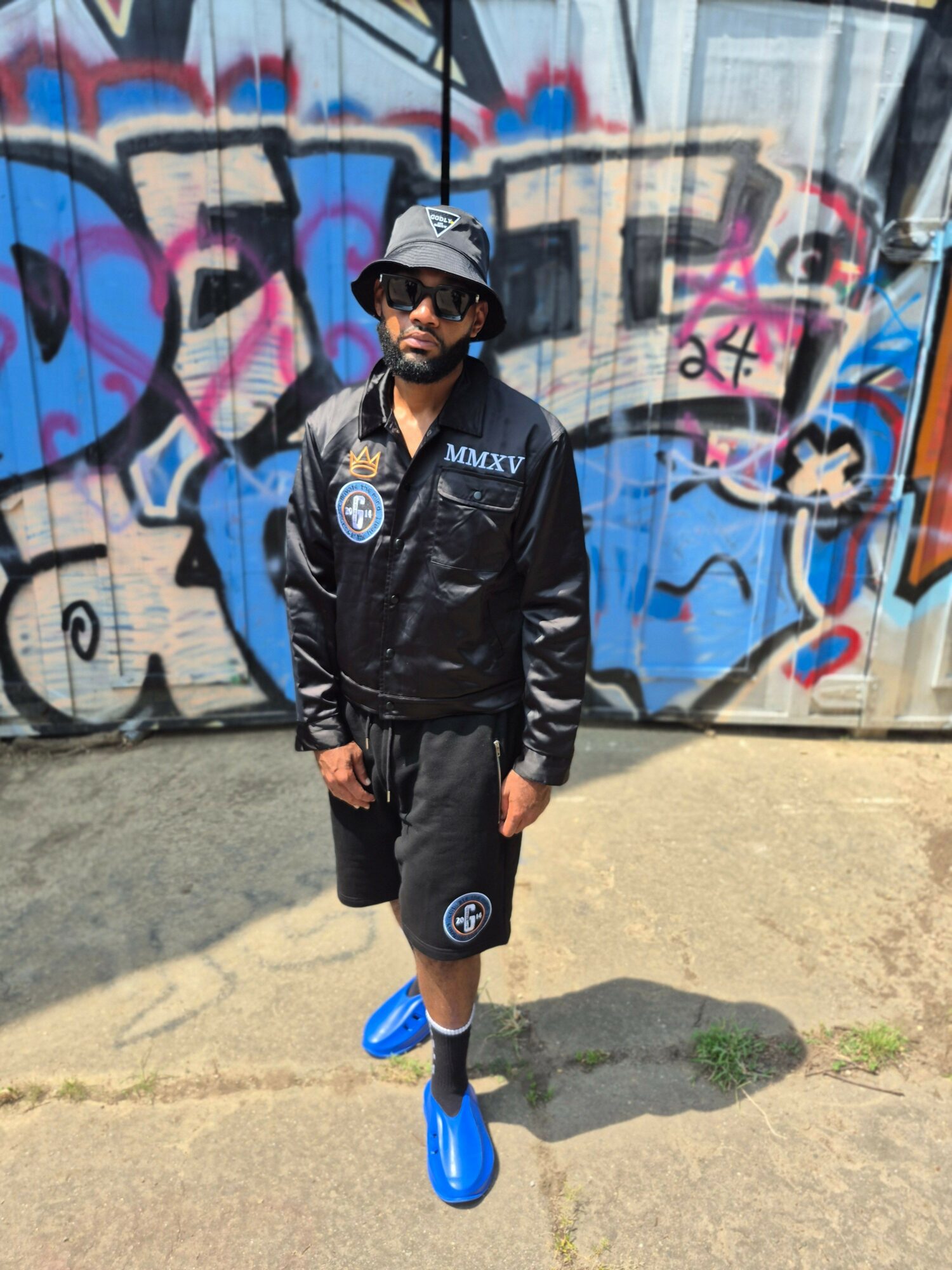 Person in black sportswear and blue shoes standing outdoors near graffiti wall, shadow cast on ground.