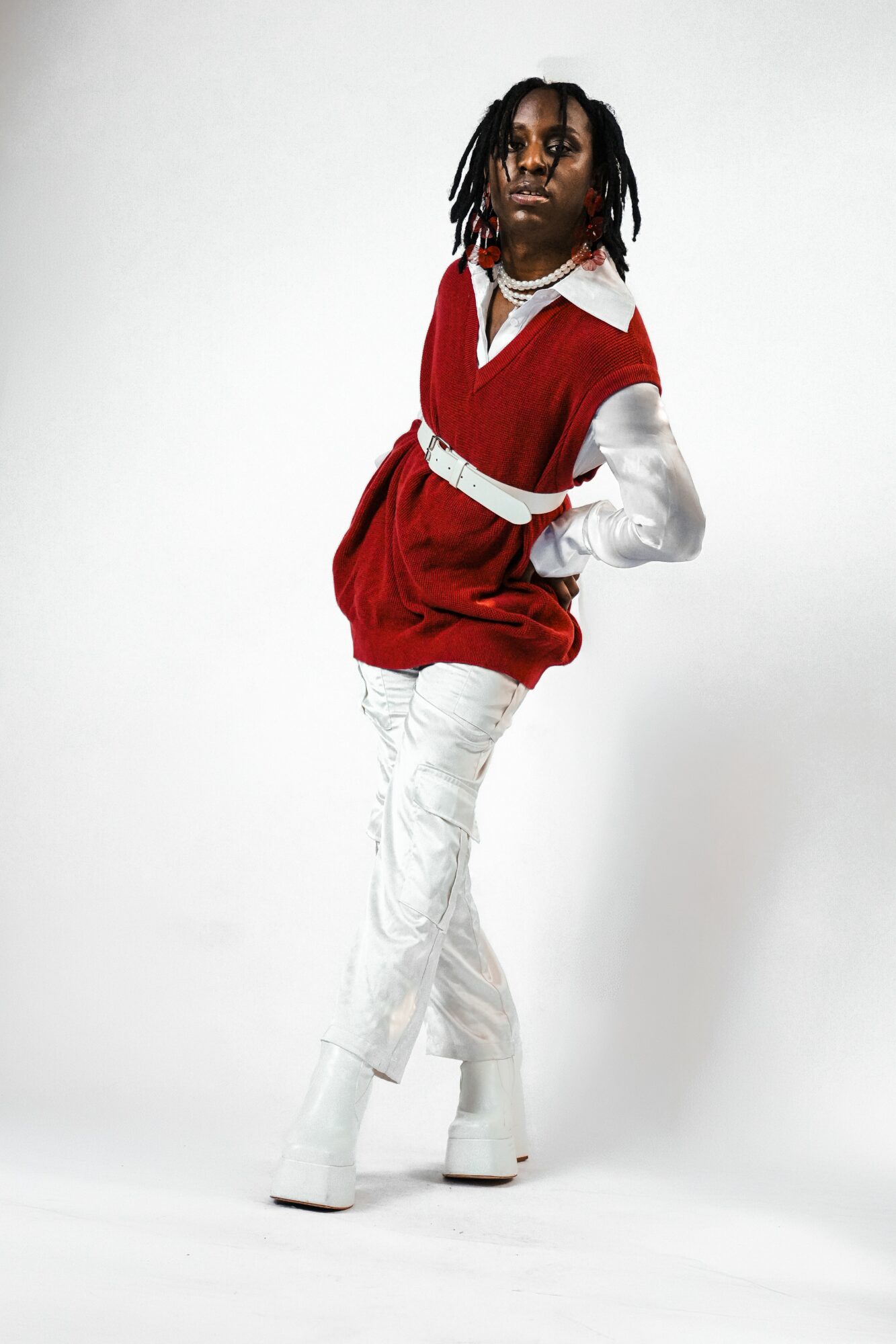 Person with dreadlocks wearing a red vest over a white shirt, white pants, and white boots, standing against a white background.