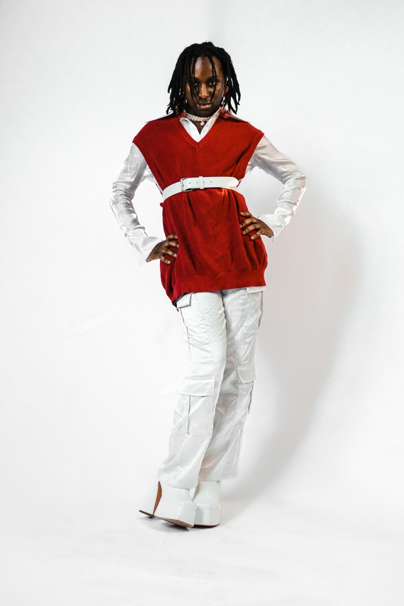 Person with dreadlocks wearing a red vest over a white shirt and white pants, standing with hands on hips against a white background.