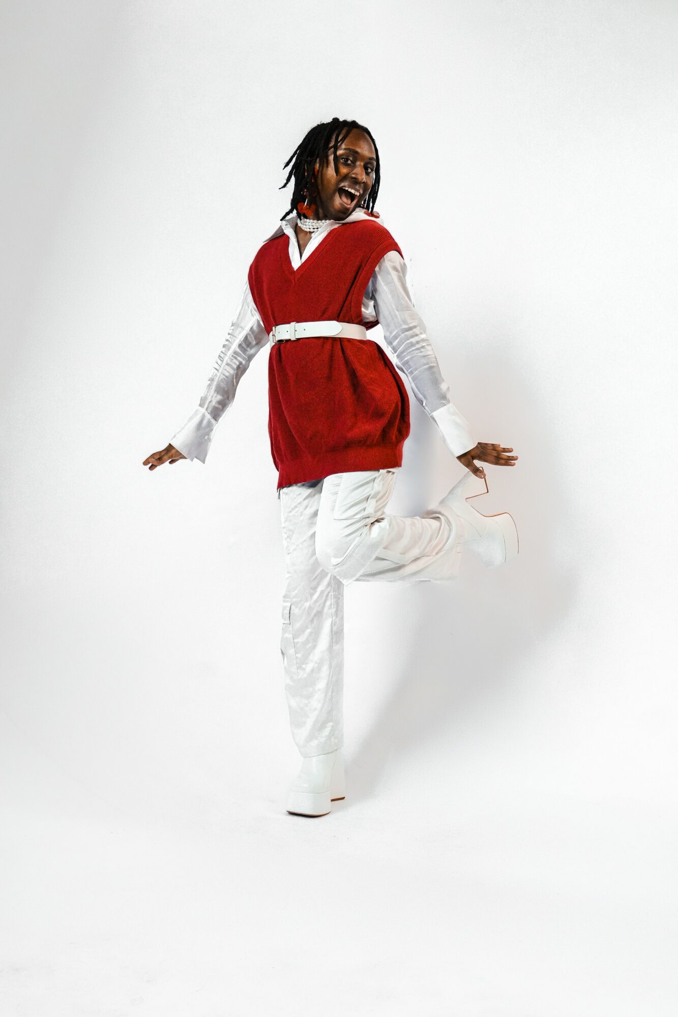 Person with dreadlocks wearing a red vest over a white shirt and white pants, smiling and jumping against a white background.