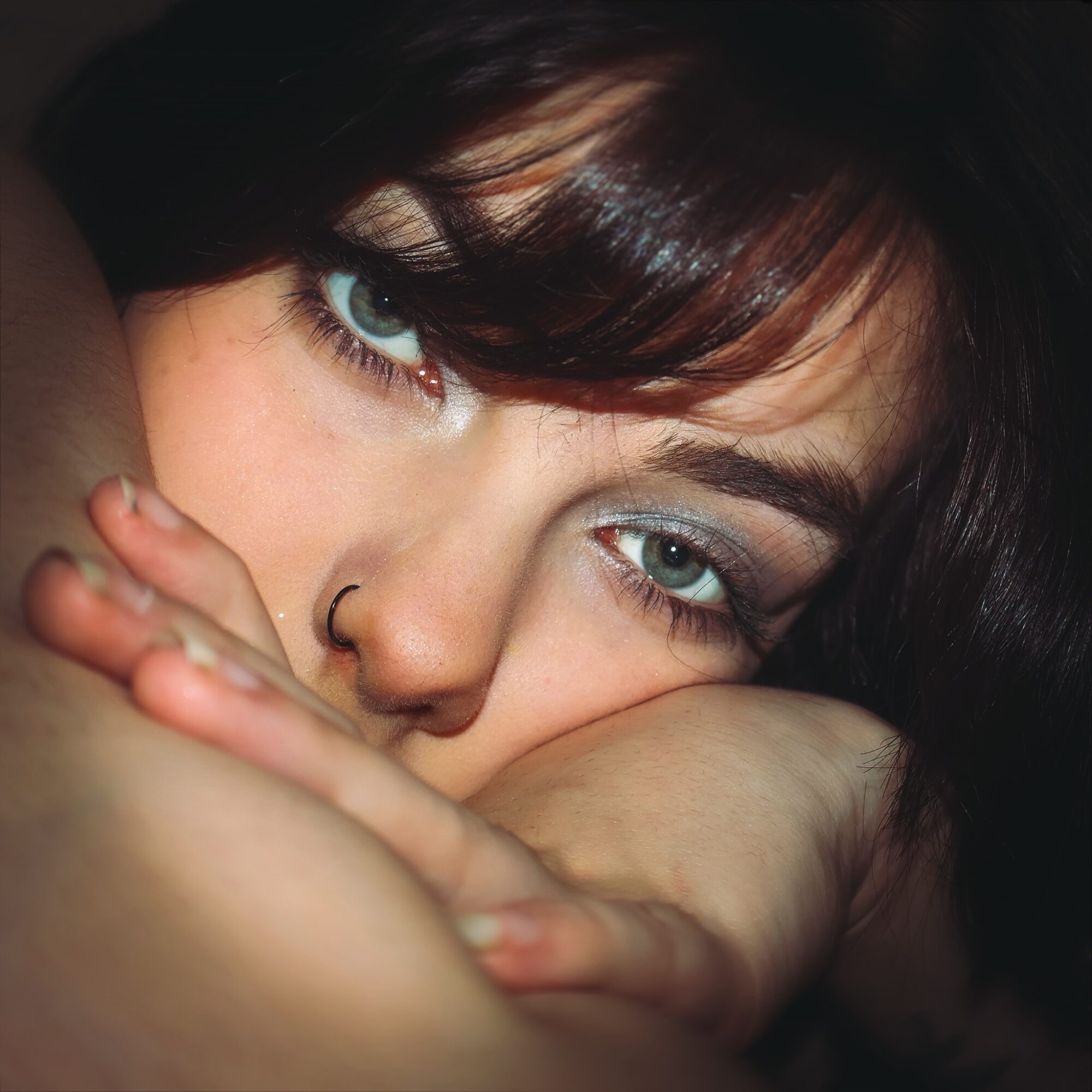 Close-up of a person with blue eyes, dark hair, and a nose ring, resting their face on their arm.