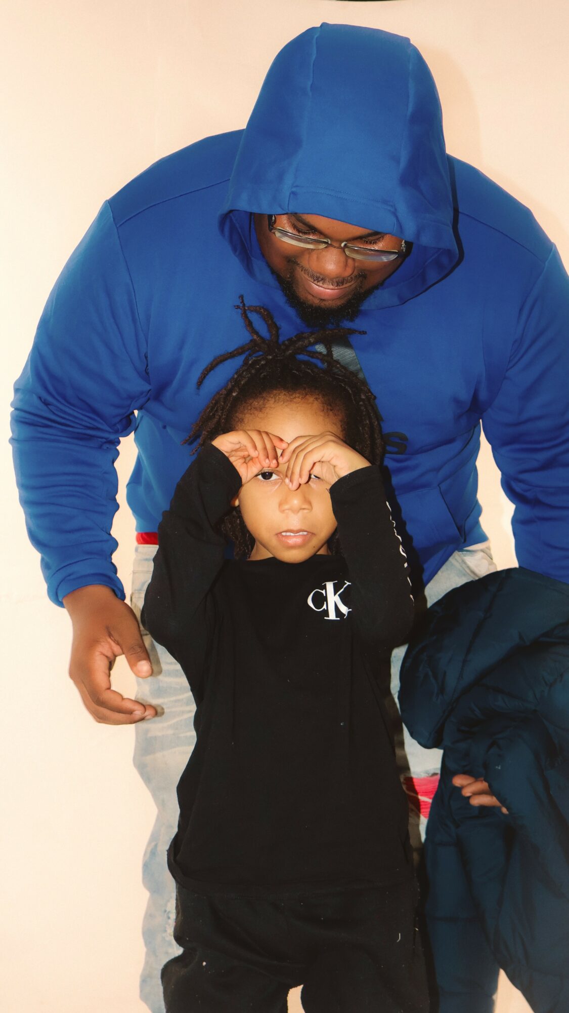 Adult and child posing together, the child making a peace sign with hands near face, both wearing dark clothing, background plain.