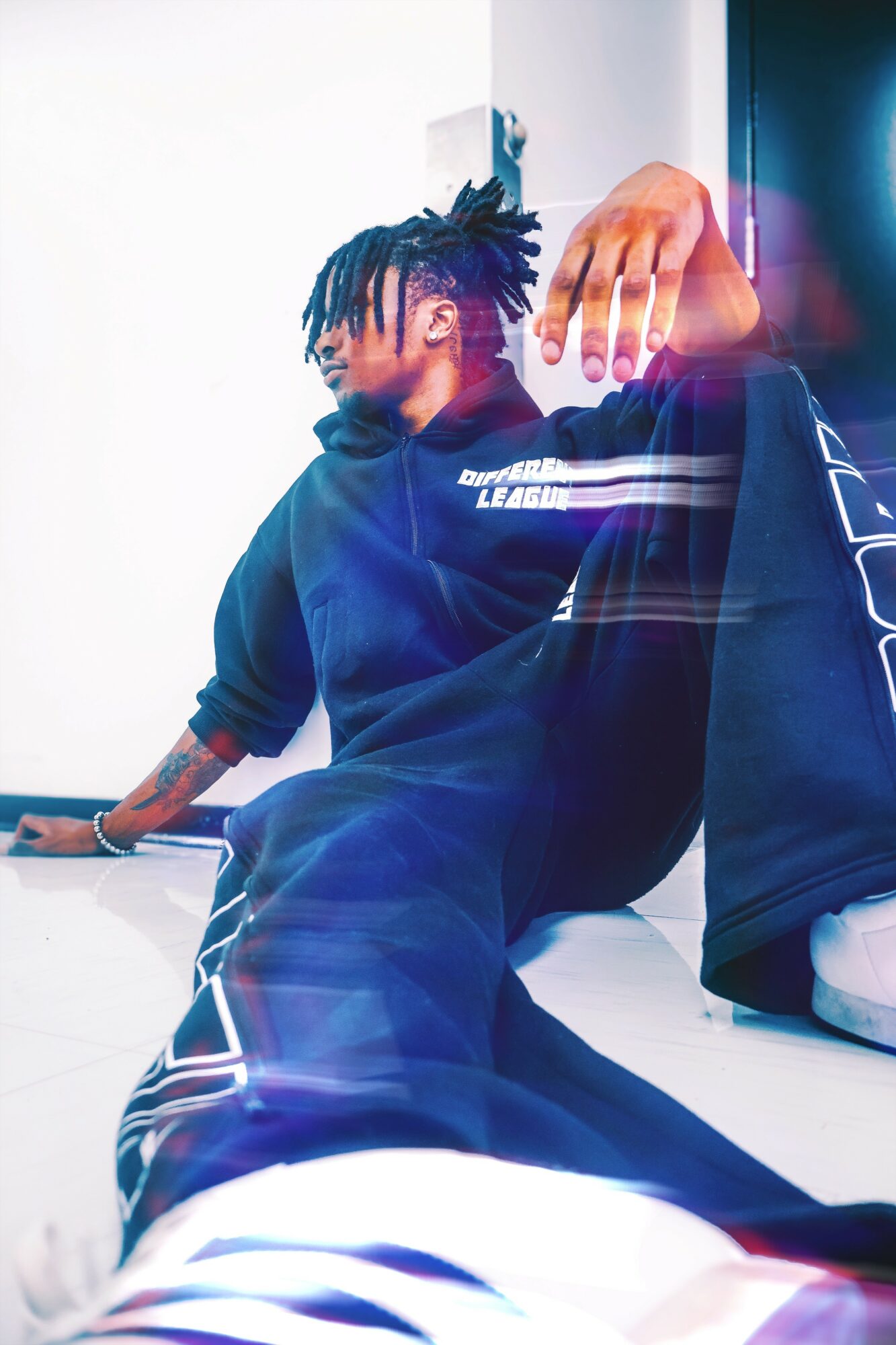 Person with dreadlocks sitting on the floor, wearing a dark hoodie and pants, in a brightly lit room.