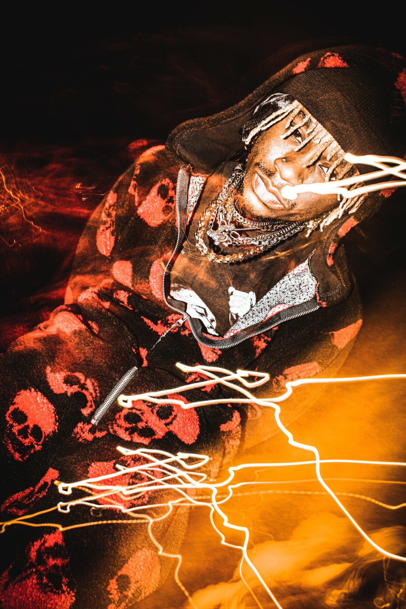Person wearing a hat and skull-patterned hoodie, with light streaks and fiery background, looking upward.