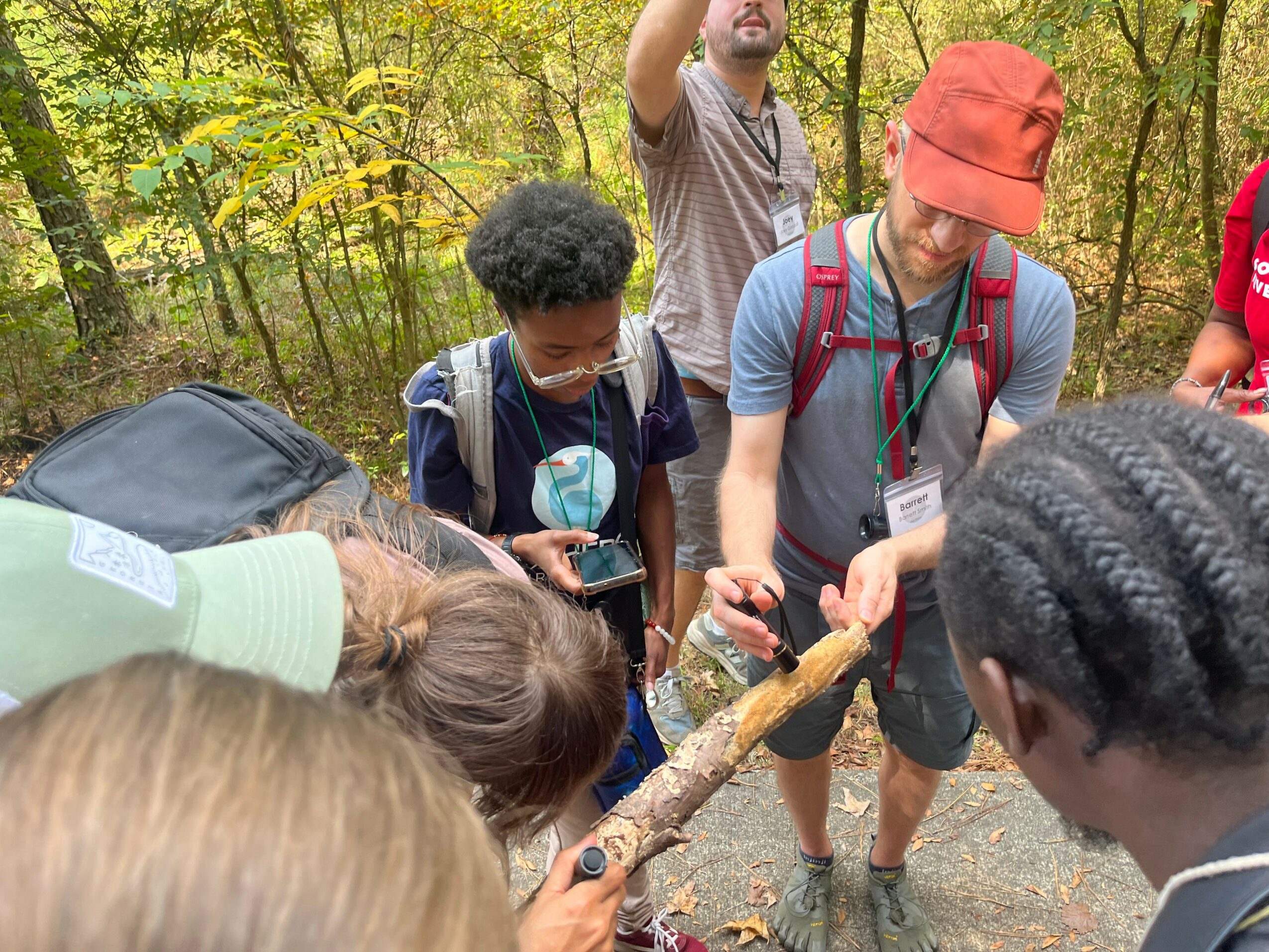 Group of people outdoors in a wooded area, some looking at a stick, others using phones, wearing backpacks and hats.