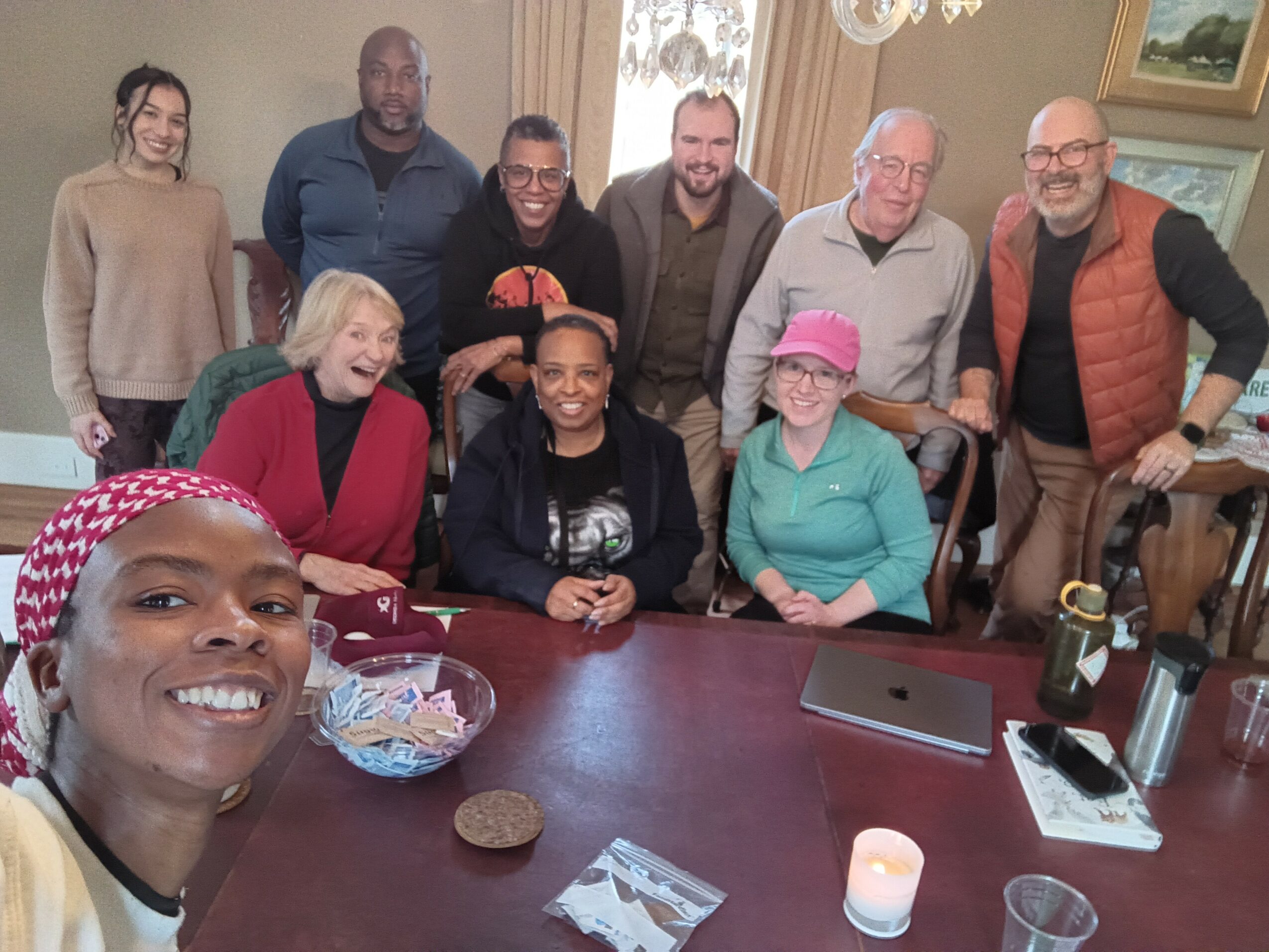 Group of ten people gathered around a table, smiling, with snacks and drinks, in a room with a window and artwork.