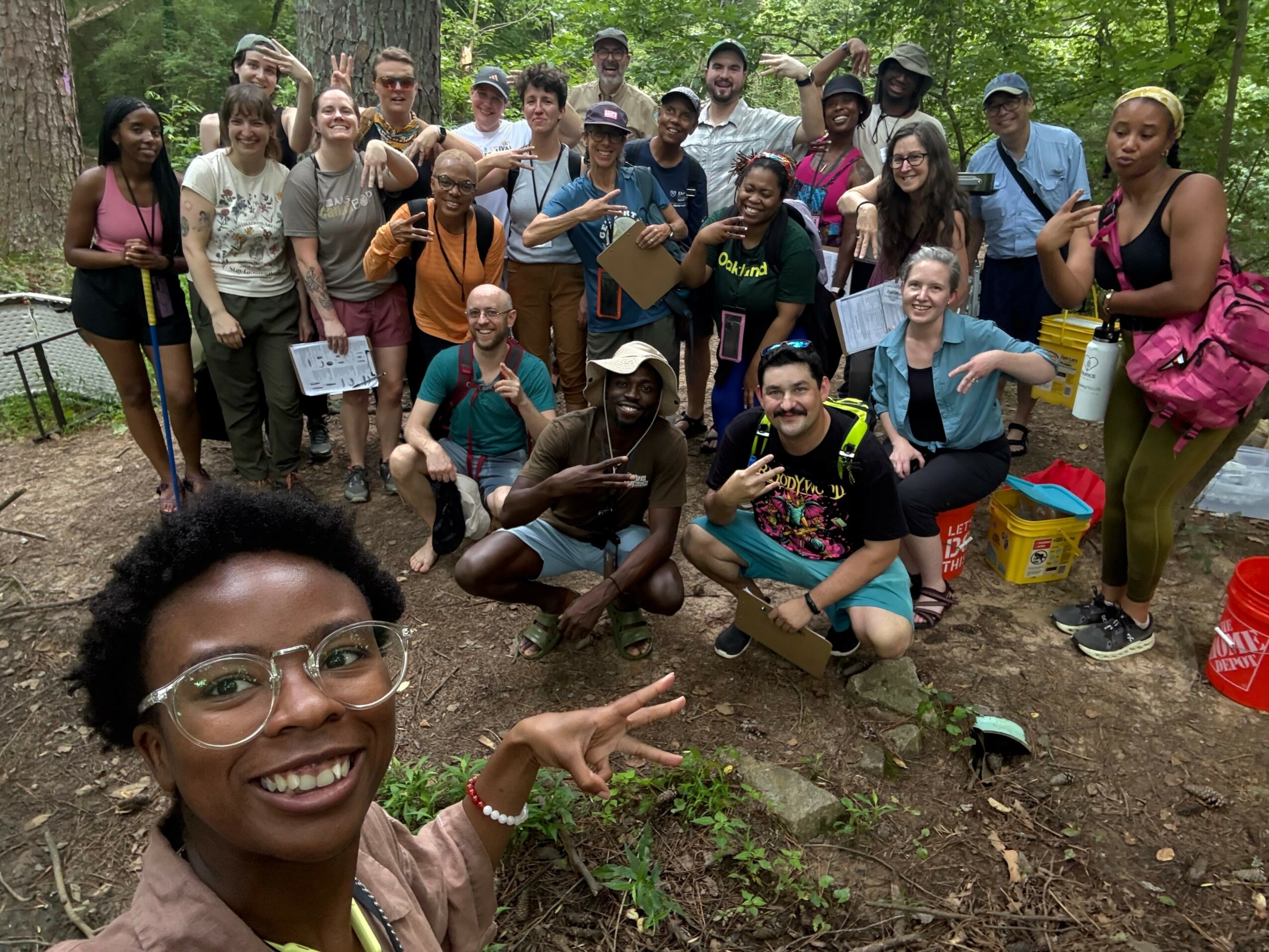 Group of diverse people outdoors in a forest, smiling and posing for a photo, some holding tools and papers.