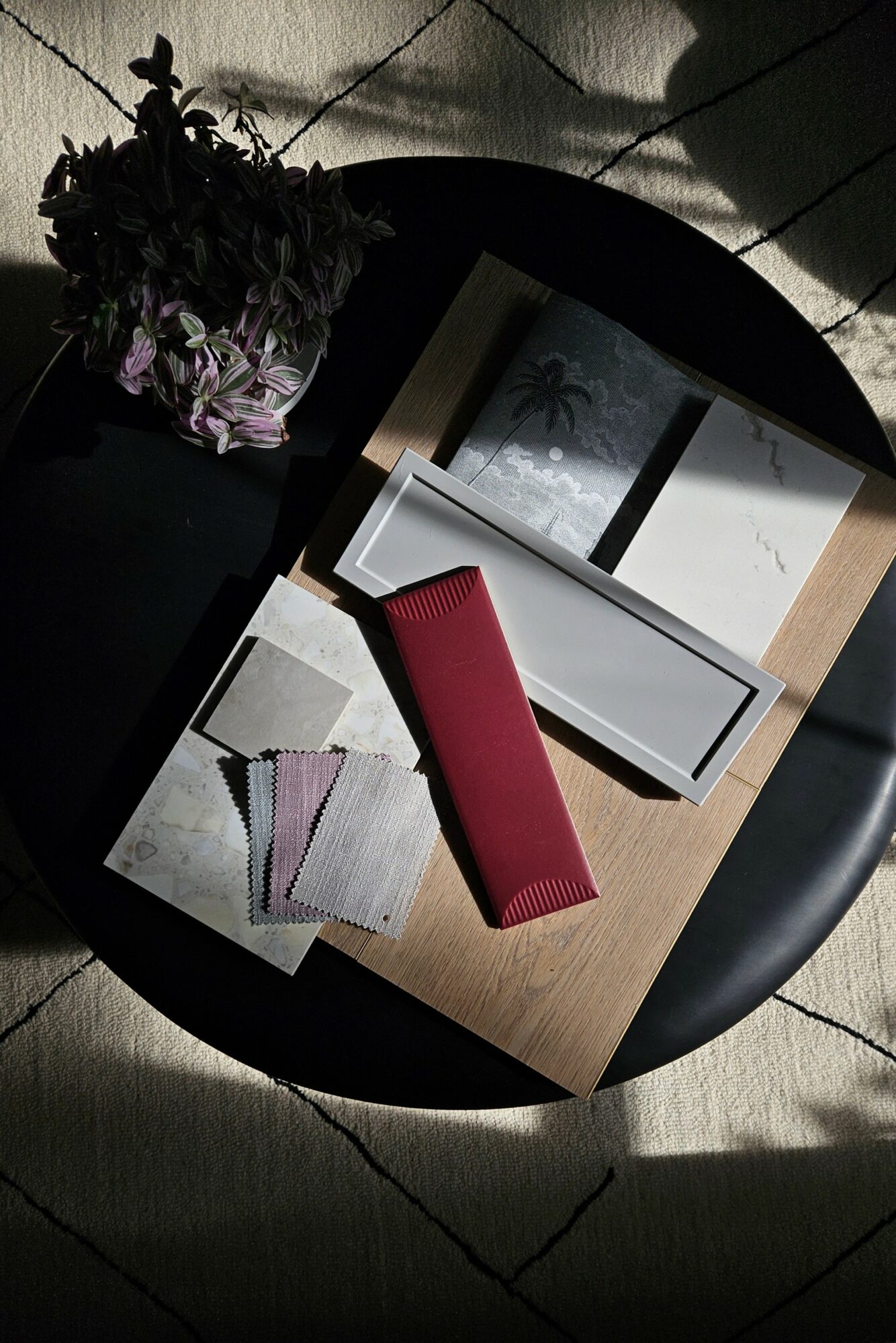 Table with books, a red case, fabric swatches, and a potted plant, illuminated by sunlight, on a tiled floor.