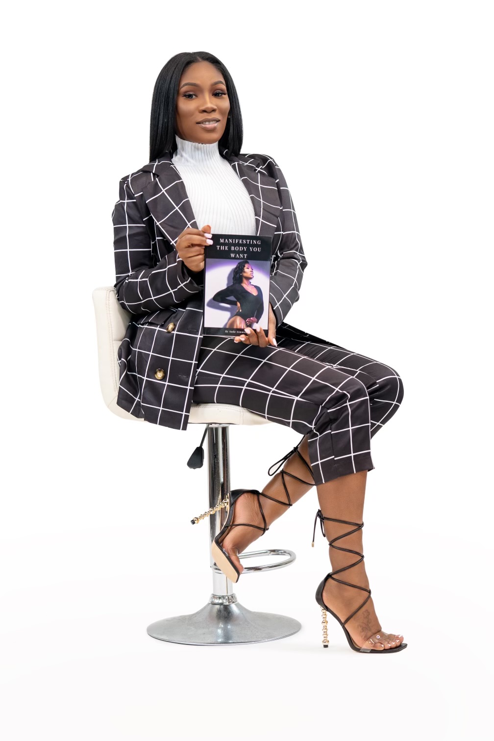 Woman sitting on a stool holding a book, wearing a checkered suit and high heels, against a white background.