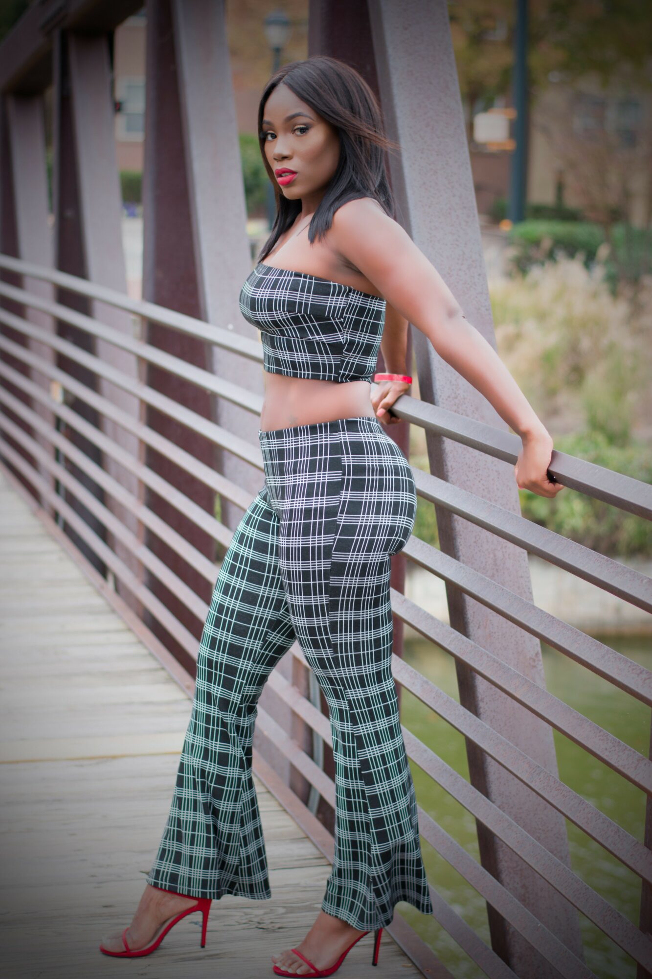 Woman in plaid outfit leaning on a railing outdoors, with trees and a building in background.