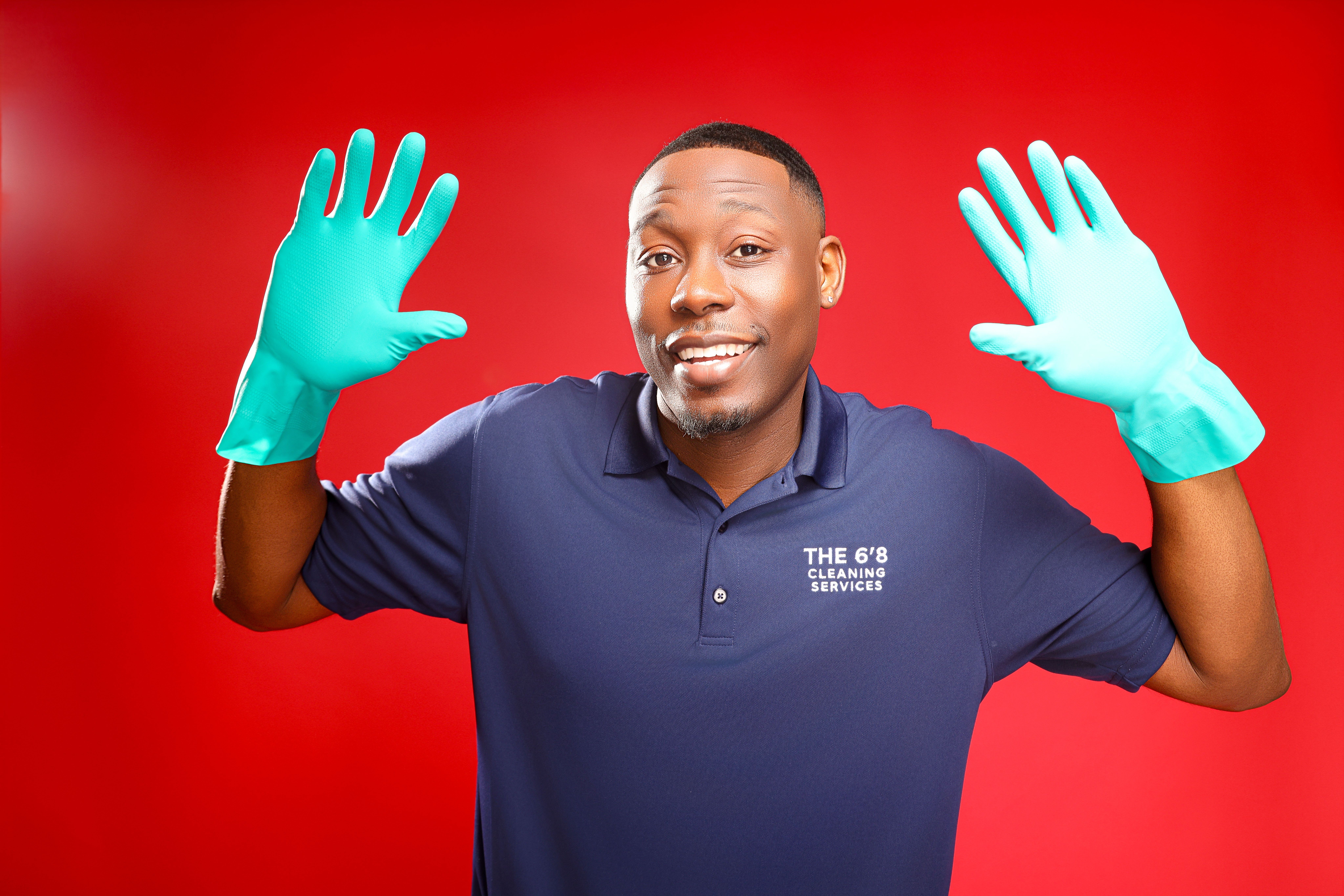 Man wearing blue shirt and teal gloves smiling with hands raised against red background.