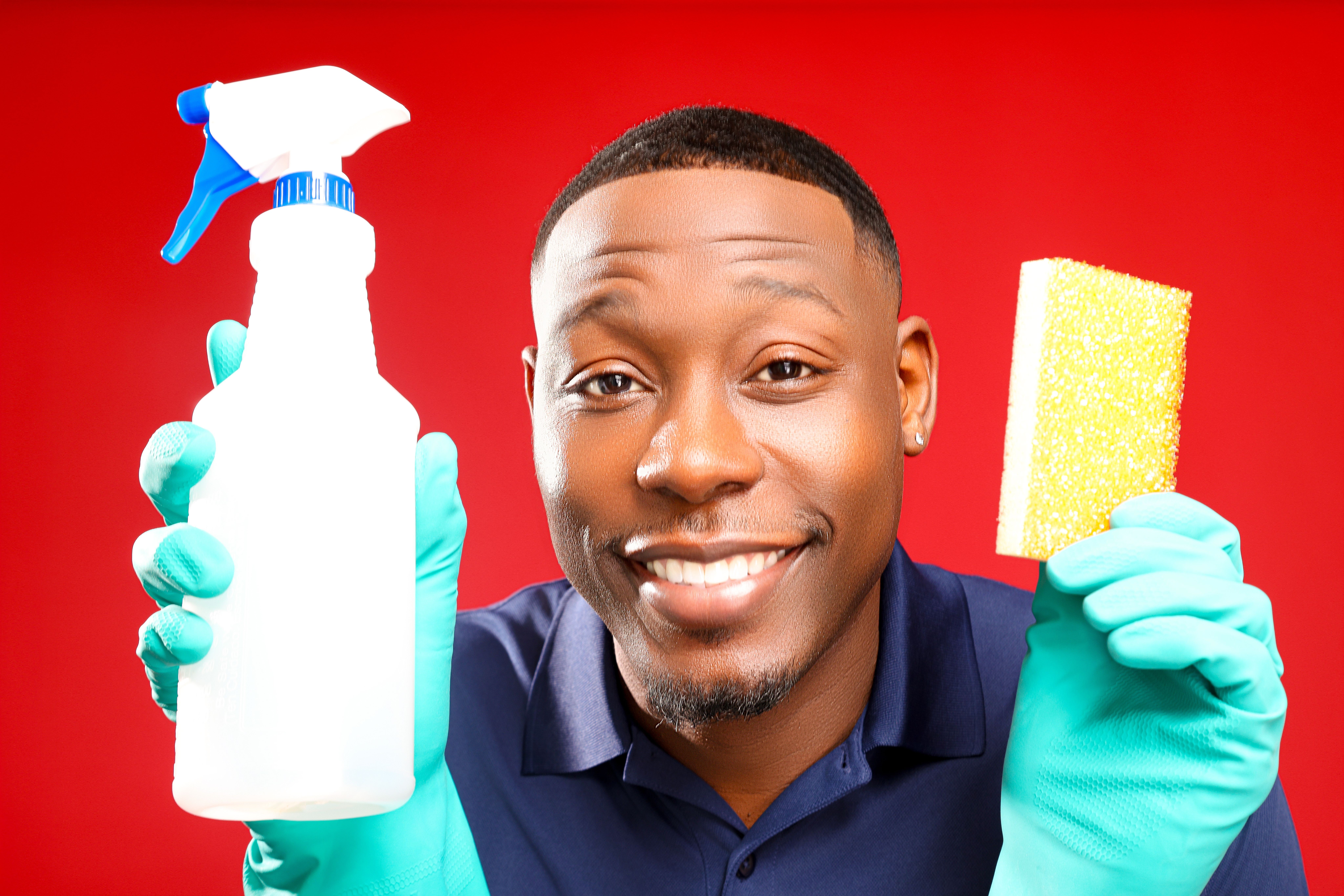 Man smiling holding cleaning spray and sponge, wearing blue gloves, against red background.
