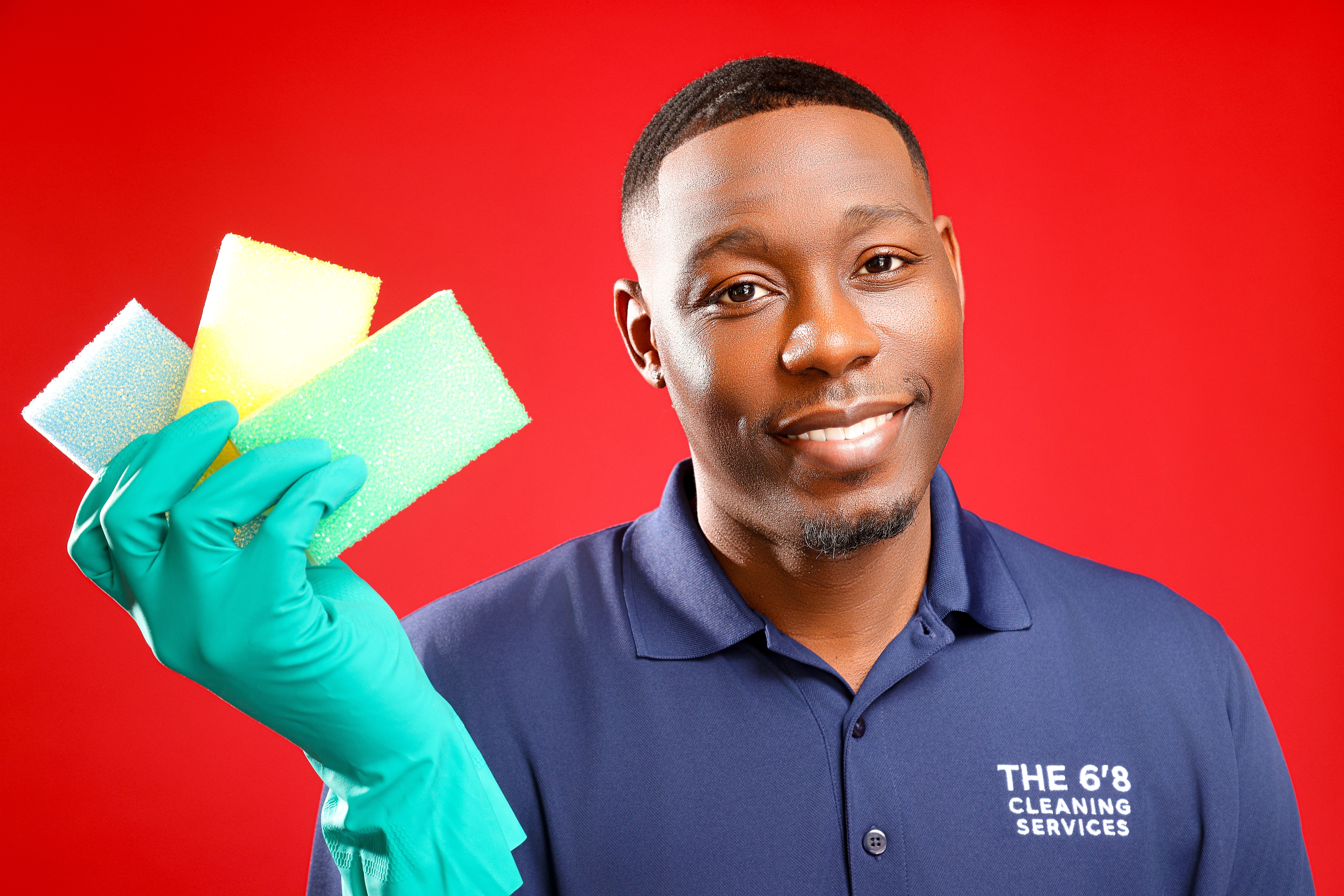 Young man wearing a blue polo shirt and green cleaning gloves holding cleaning sponges, smiling against a red background.