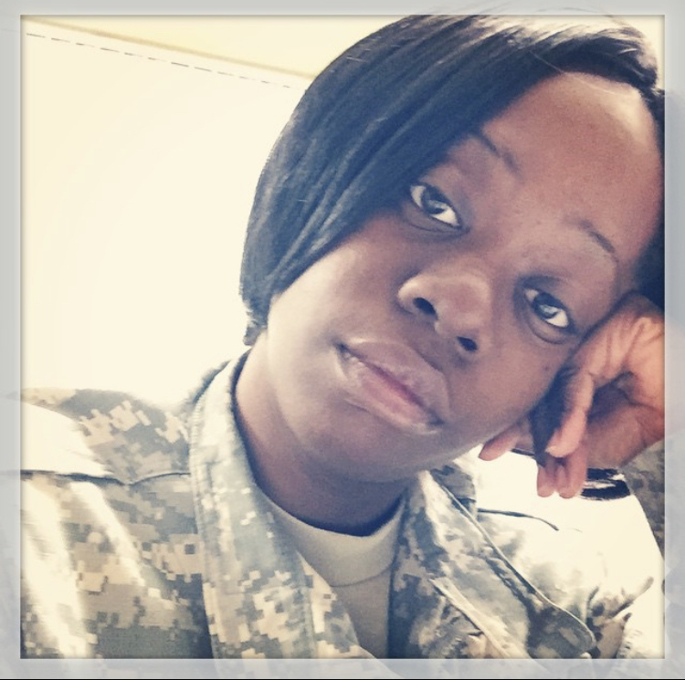 Person in military uniform resting head on hand, looking at camera, indoors, with light background.