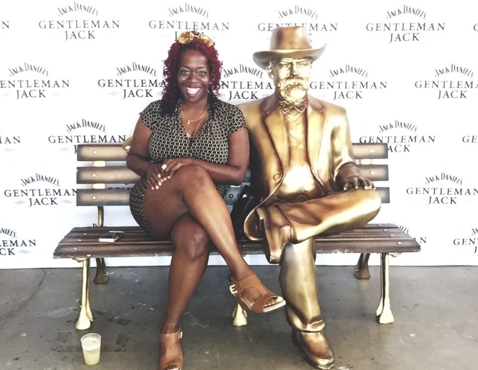 Woman sitting on a bench next to a golden statue of a man in a hat, both smiling, backdrop with 'Gentleman Jack' logo.