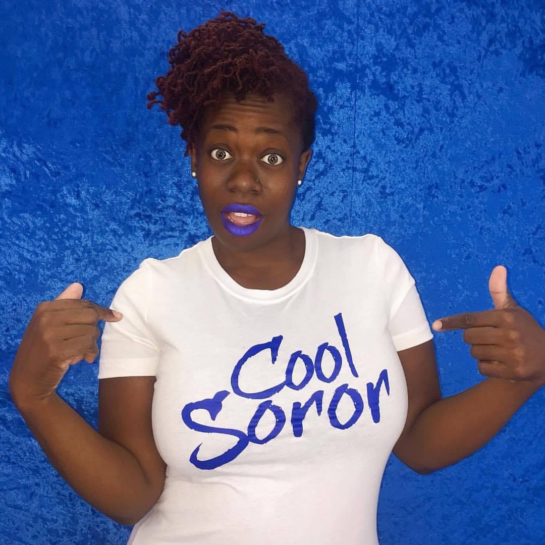 Woman with curly hair pointing to her shirt, which says 'Cool Soror', against a blue background.