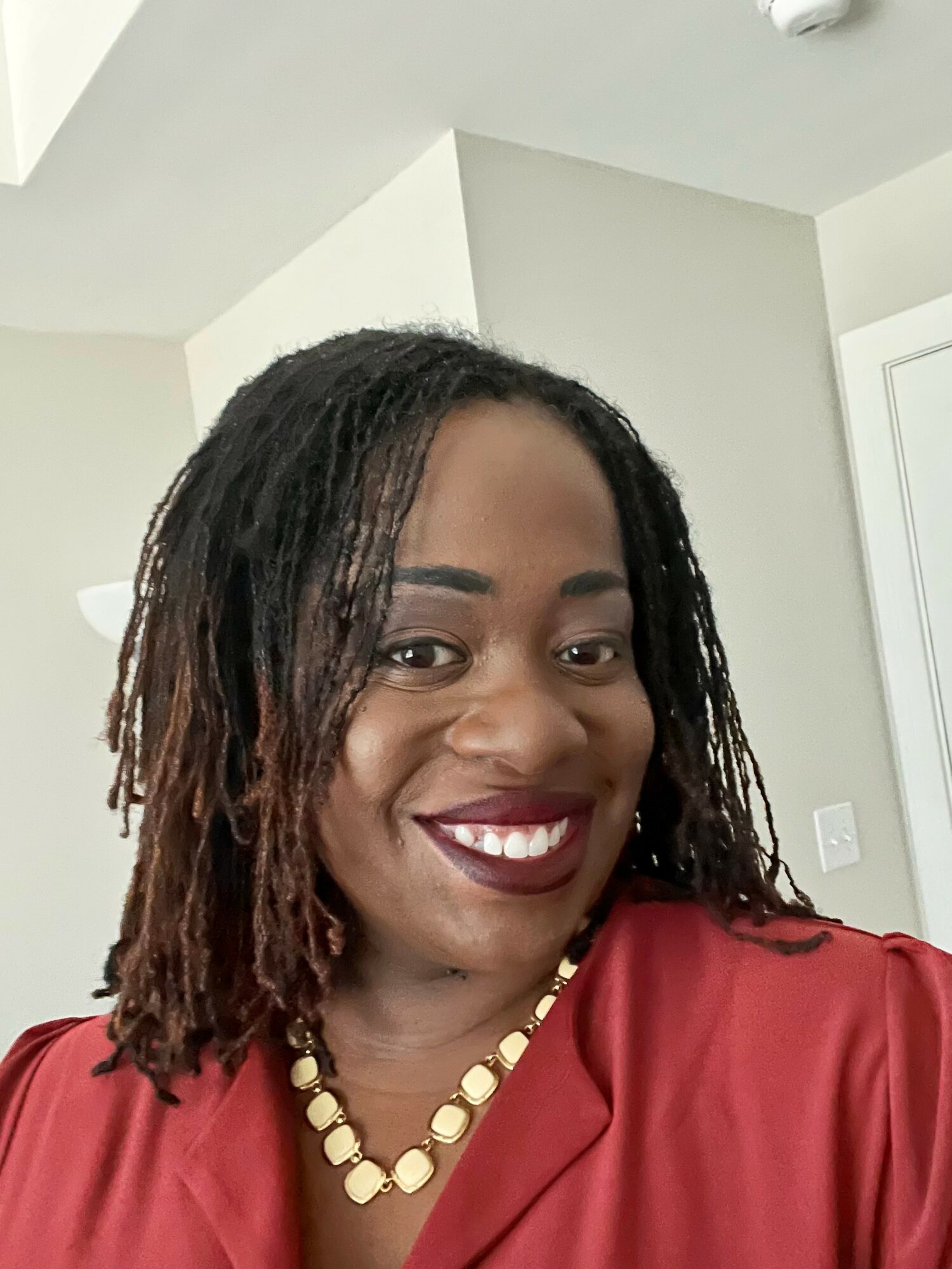 Smiling woman with shoulder-length dreadlocks wearing a red top and a gold necklace.