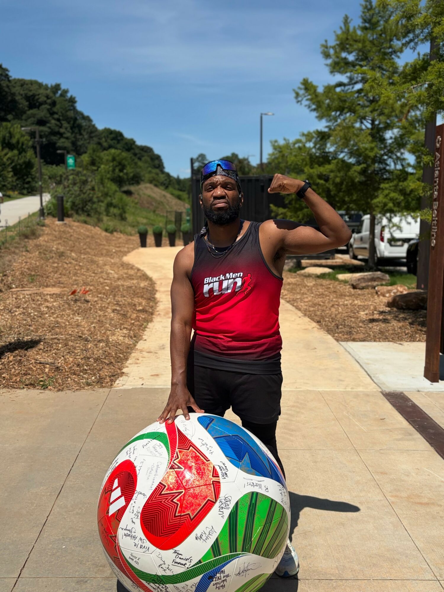 Man standing outdoors on sidewalk, holding a large colorful inflatable ball, wearing a red tank top and sunglasses.
