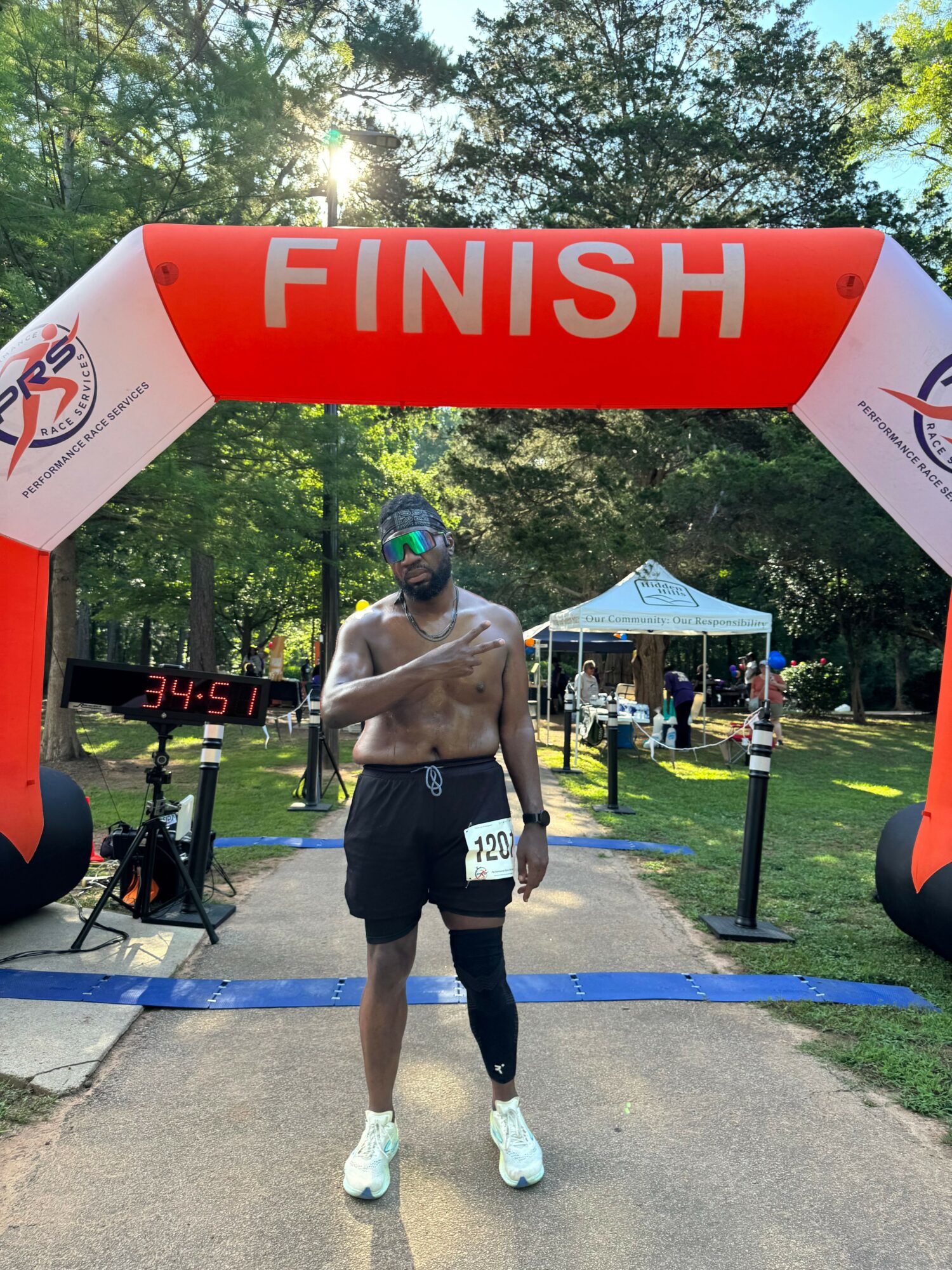 Runner crossing finish line with a red and white arch, trees and a tent in background, person wearing black shorts and white shoes