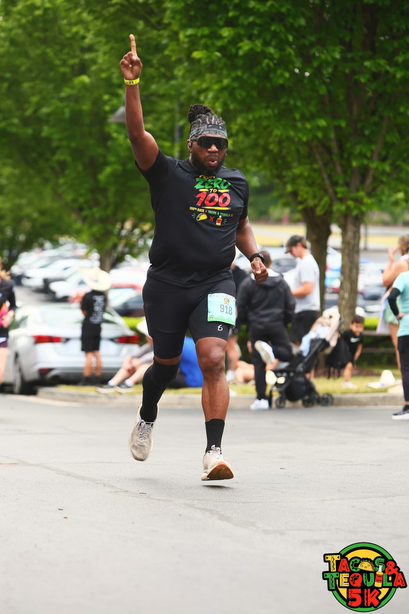 Runner with a headband and sunglasses raising one arm during a race, with trees and spectators in background.