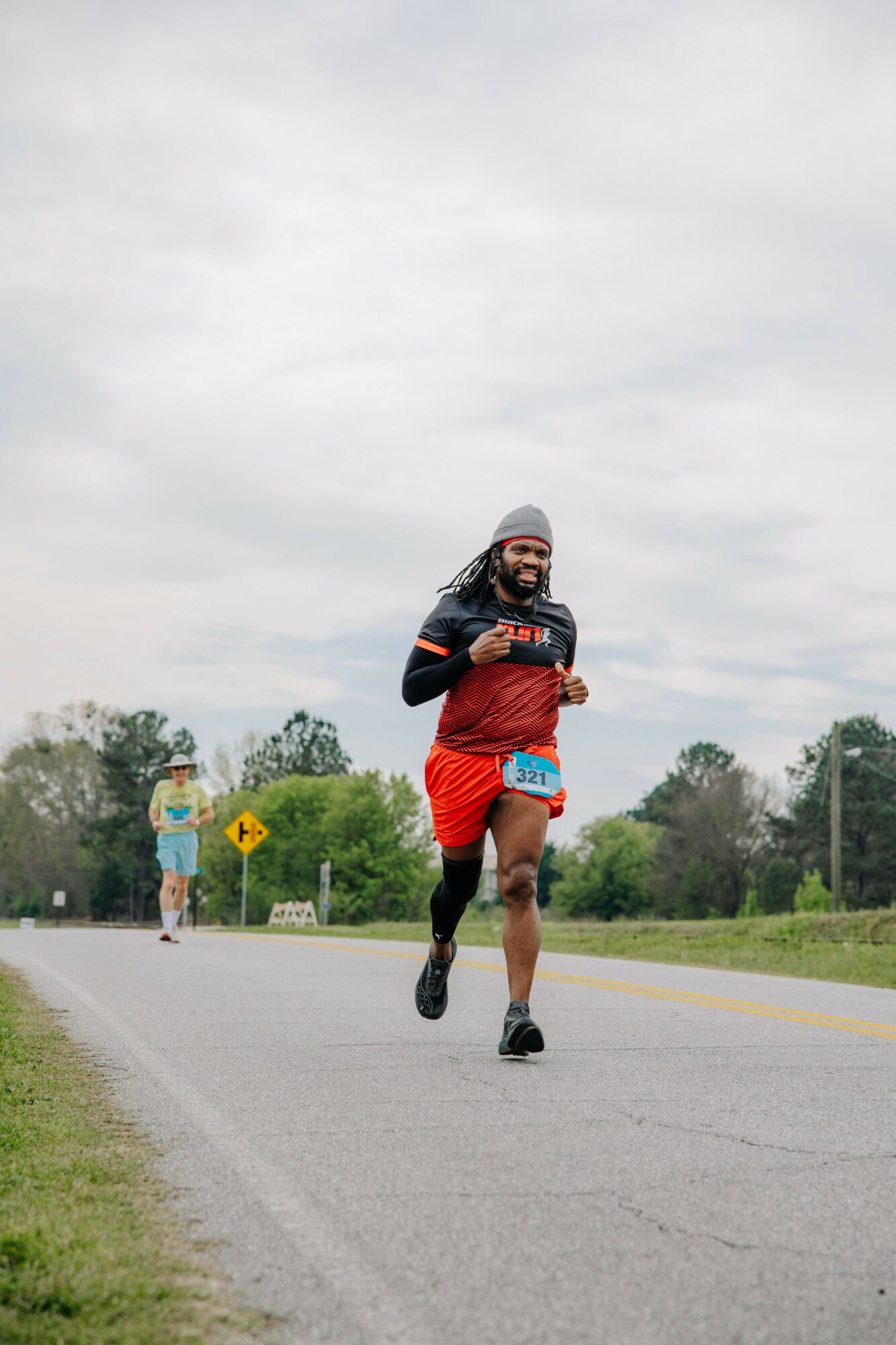 Person running on road wearing red and black athletic clothing, gray hat, and sunglasses, with trees and cloudy sky in background.