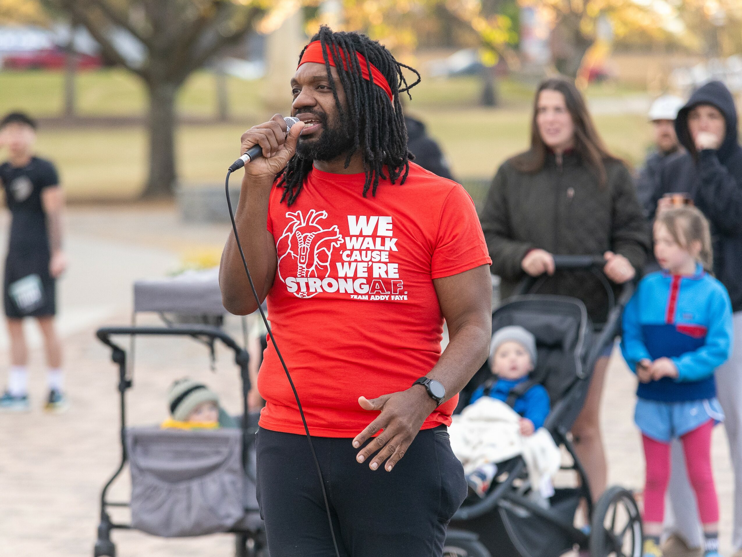 Man with dreadlocks speaking into microphone outdoors, wearing red T-shirt, surrounded by people and strollers, trees in background.
