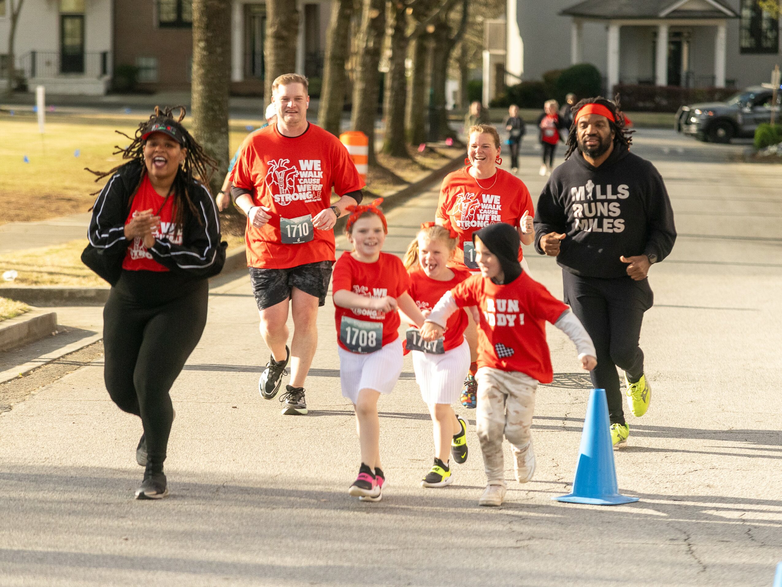 Group of people, including children and adults, running outdoors on a sunny day, some wearing red shirts, with trees and houses in background.