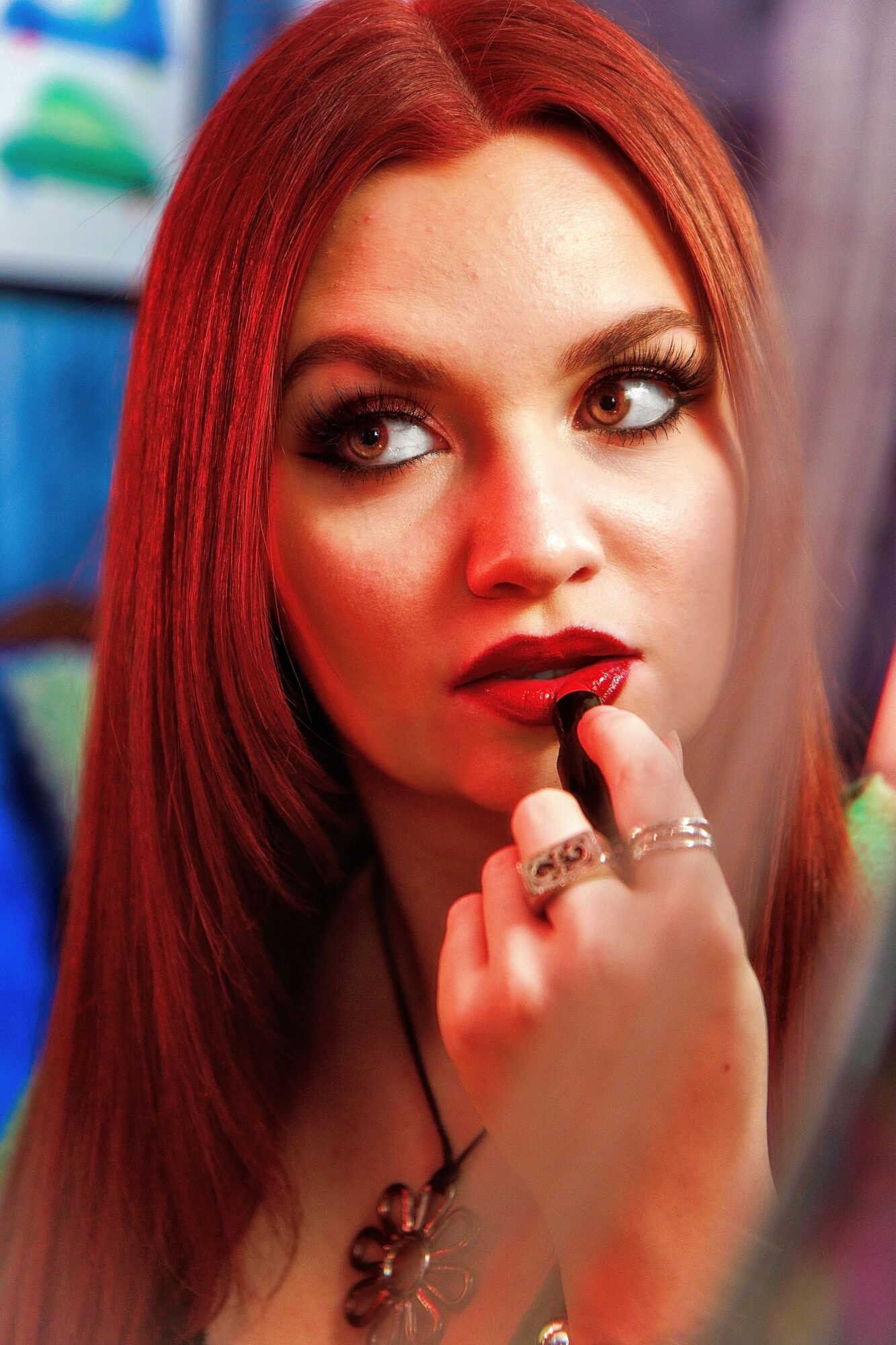 Young woman with red hair applying lipstick, looking to the side, in a colorful background.
