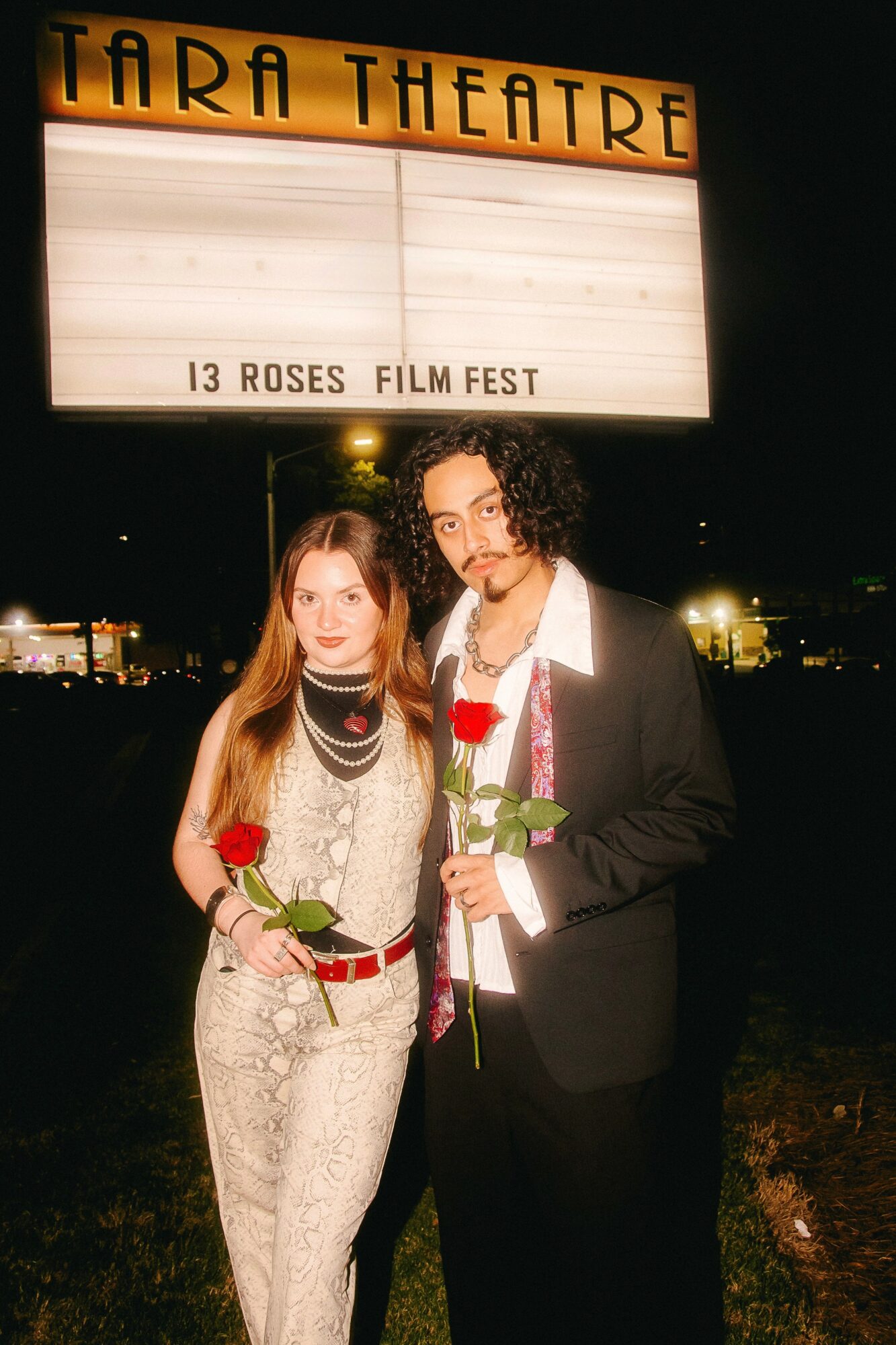 Two people holding roses stand in front of a theater sign at night, with the sign reading 'Tara Theatre' and event details.