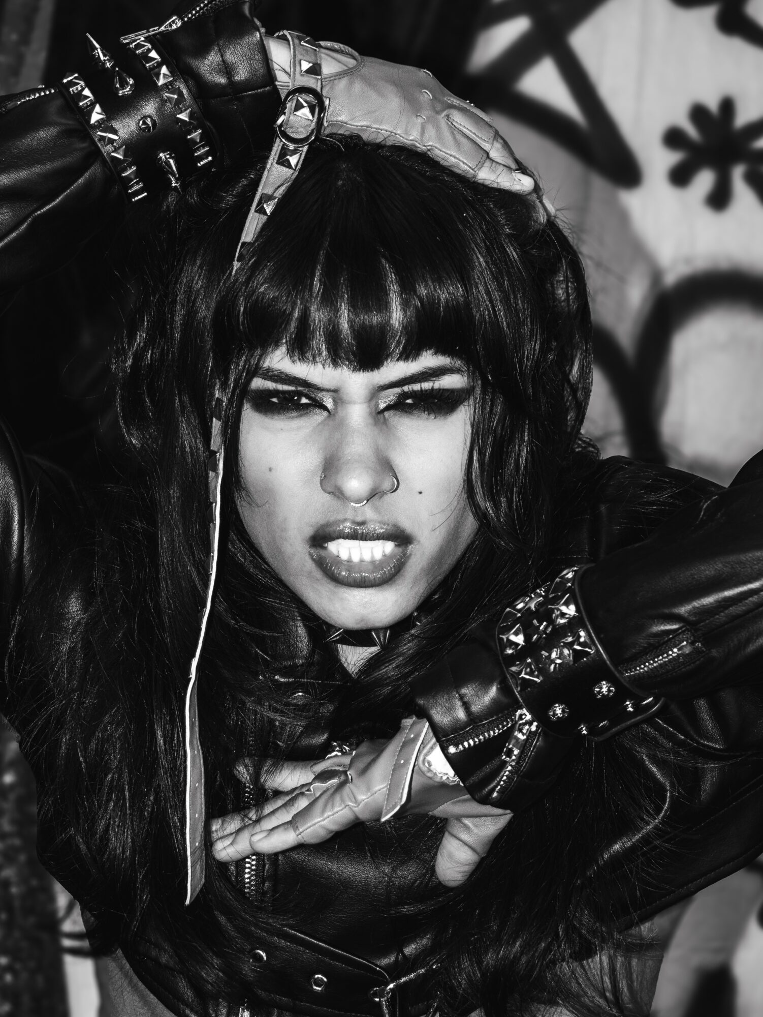 Woman with dark hair and bangs, making a fierce facial expression, wearing leather gloves and accessories, graffiti background.
