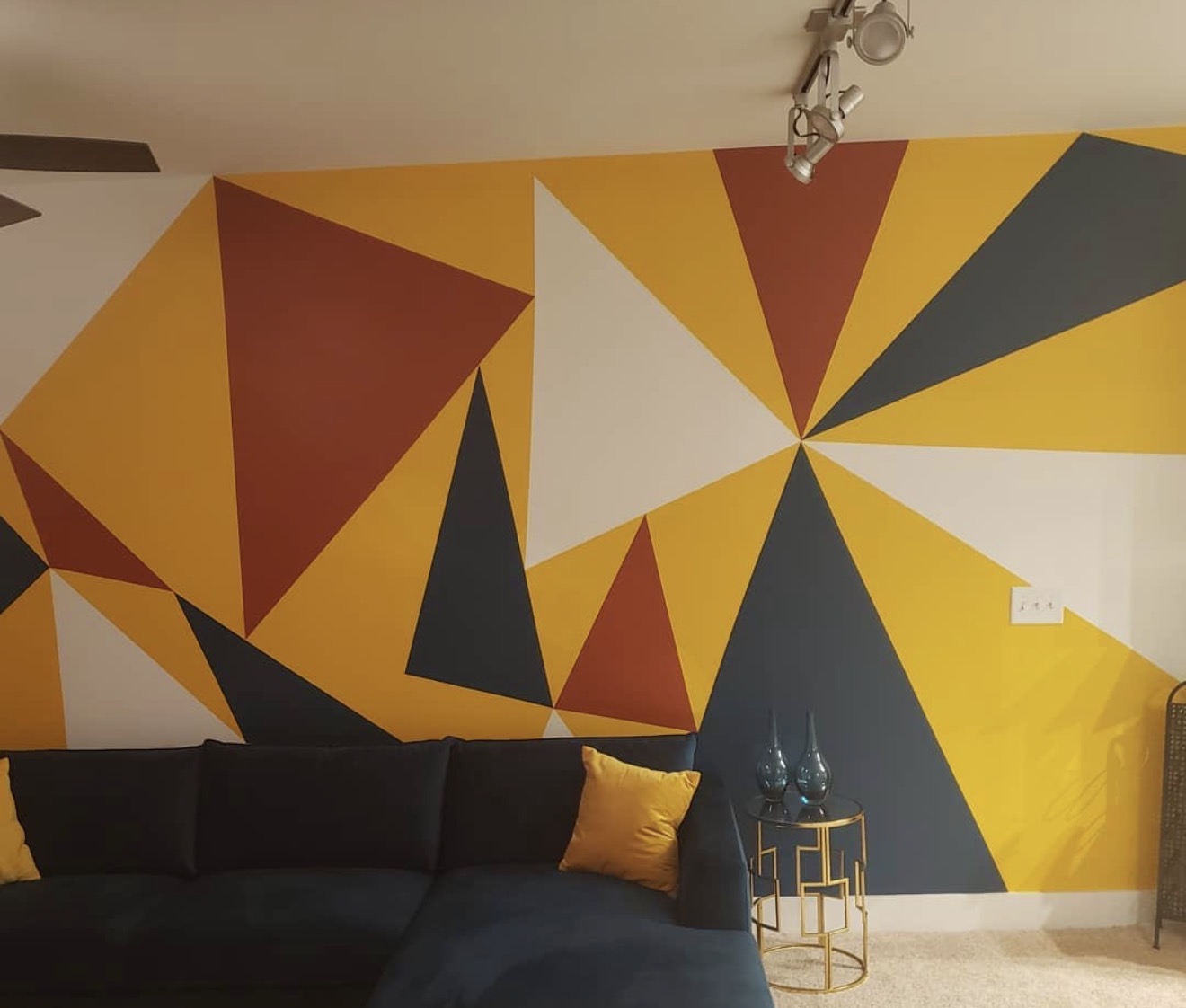 Colorful geometric mural on wall with black sofa and small side table, yellow pillow, and ceiling light fixture.