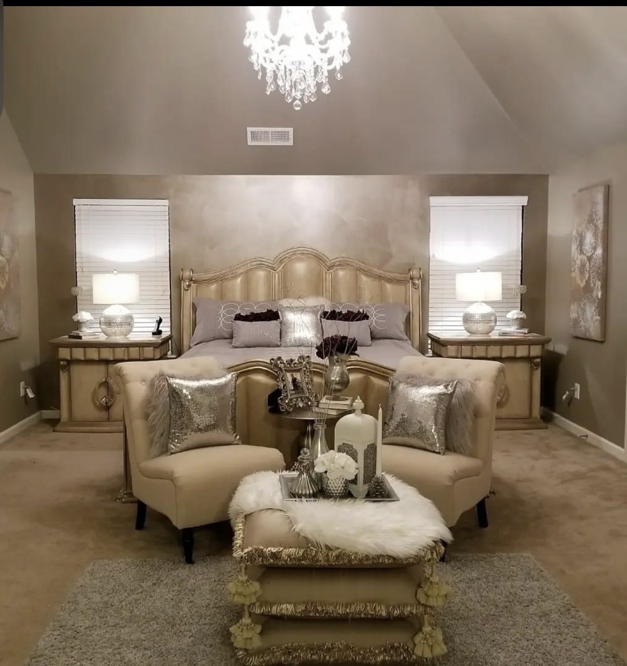 Elegant bedroom with a large bed, two nightstands with lamps, a chandelier, and a sitting area with pillows and decorative items.