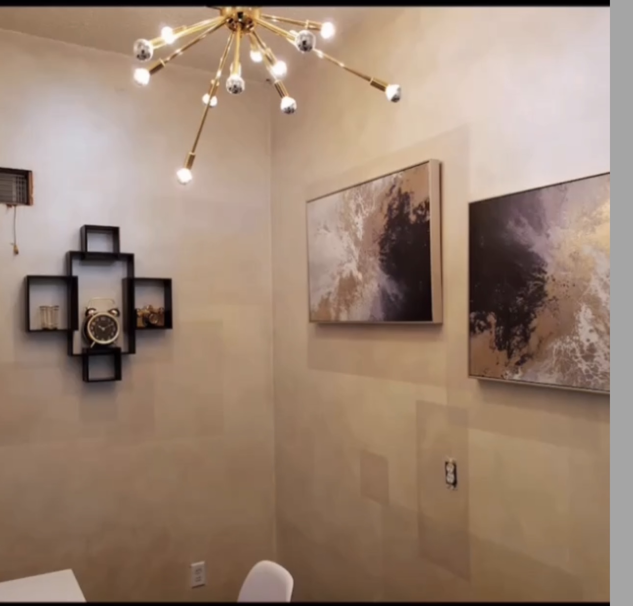 Corner of a room with wall art, a modern chandelier, and a wall clock, with a chair partially visible.