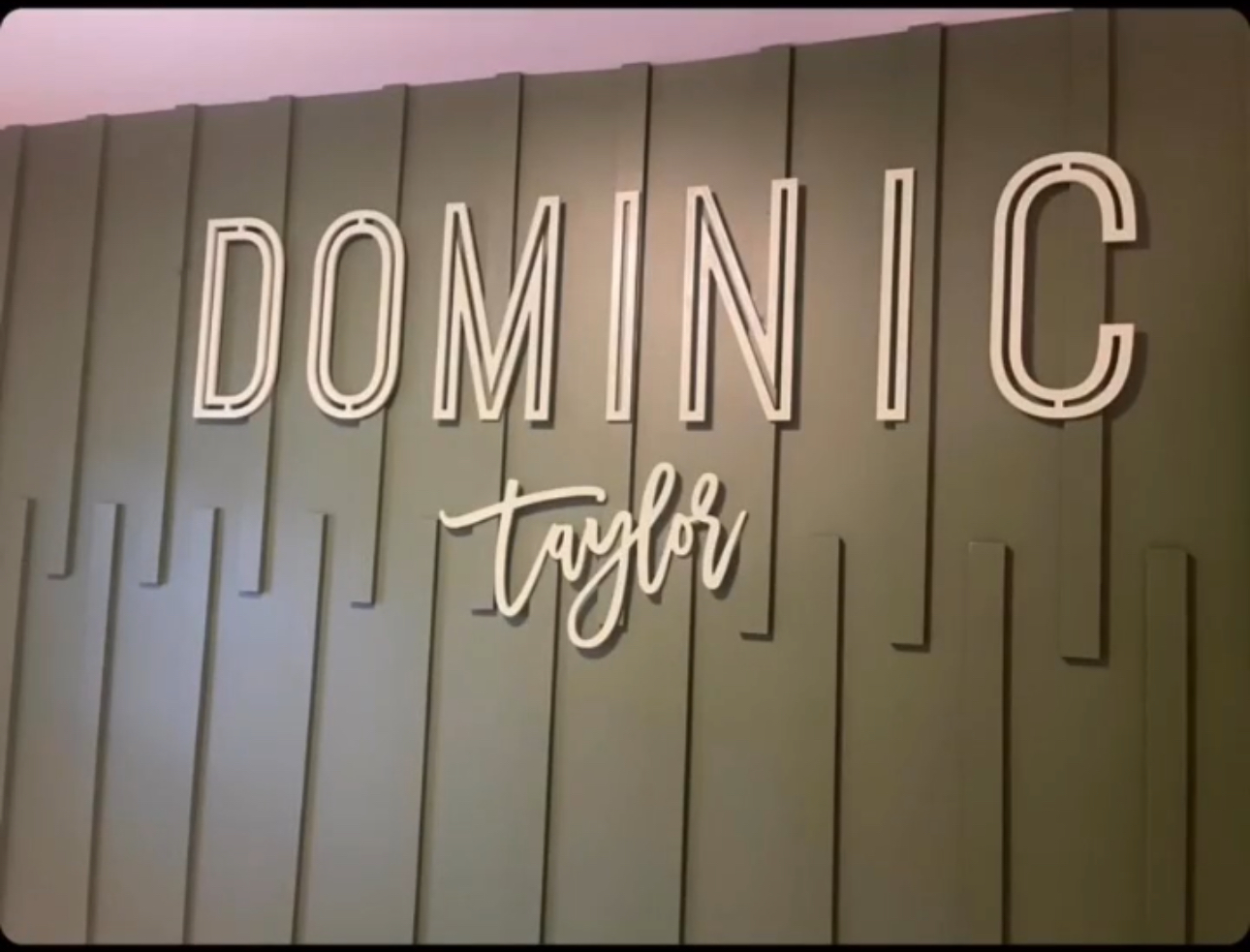 Sign with large illuminated letters reading 'DOMINIC' and smaller cursive 'table' on a wall with vertical panels.