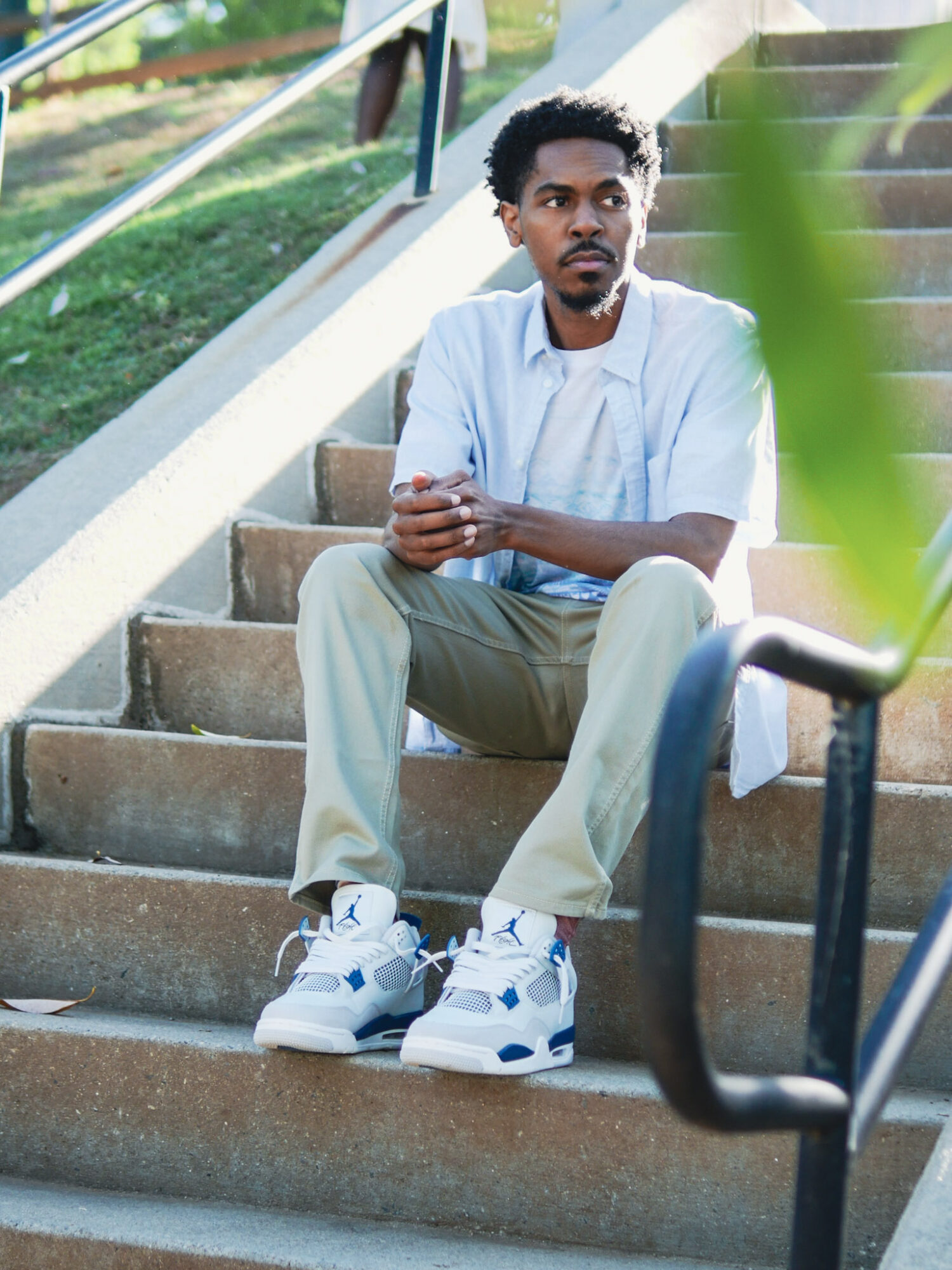 Young man sitting on outdoor stairs with greenery in background, wearing light-colored clothing and sneakers, looking to the side.