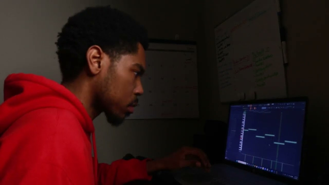 Young man in red hoodie working on a computer in a dark room, with a whiteboard and monitor in the background.