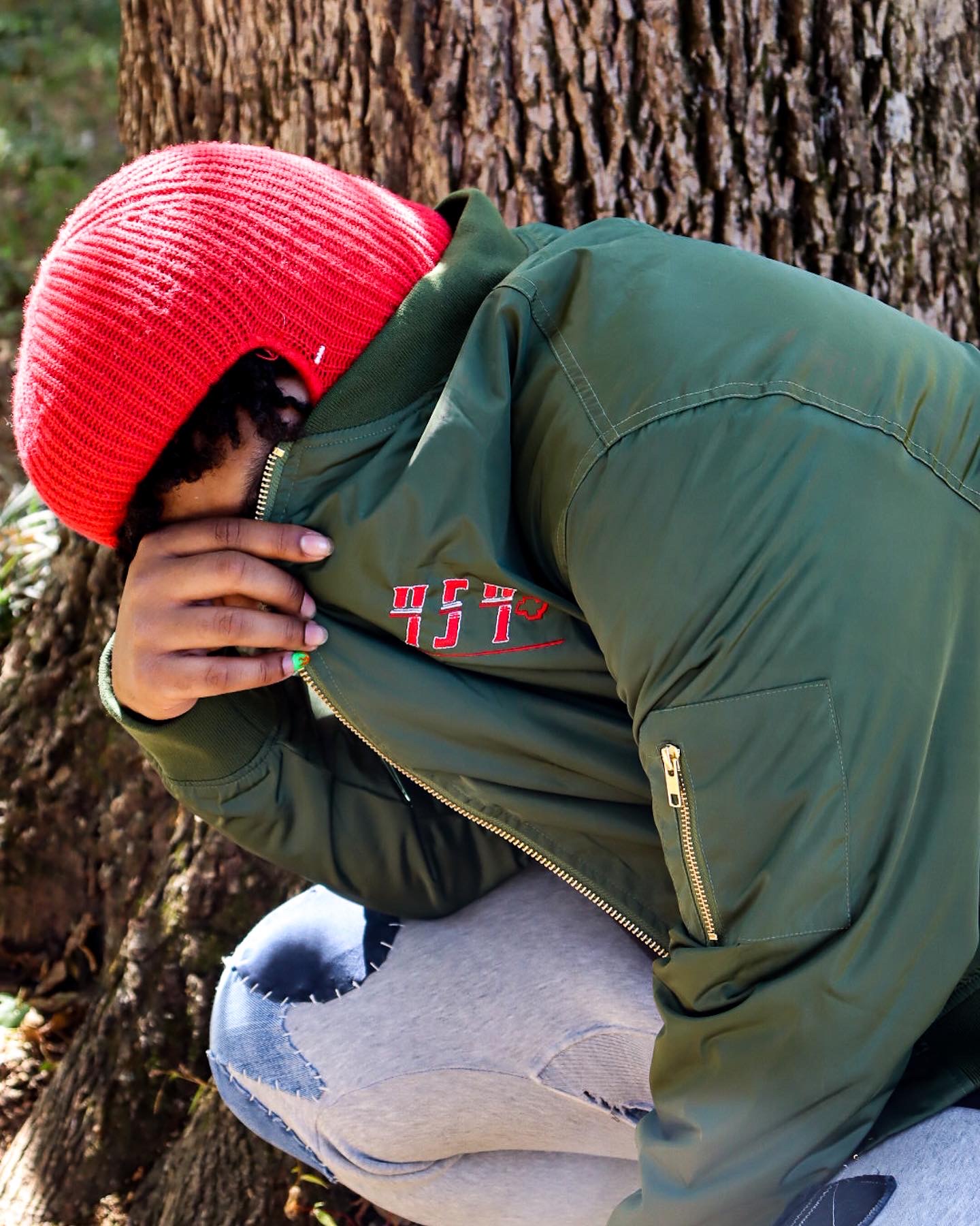 Person wearing a red beanie and green jacket, crouching near a tree outdoors, with hand near face, in natural setting.