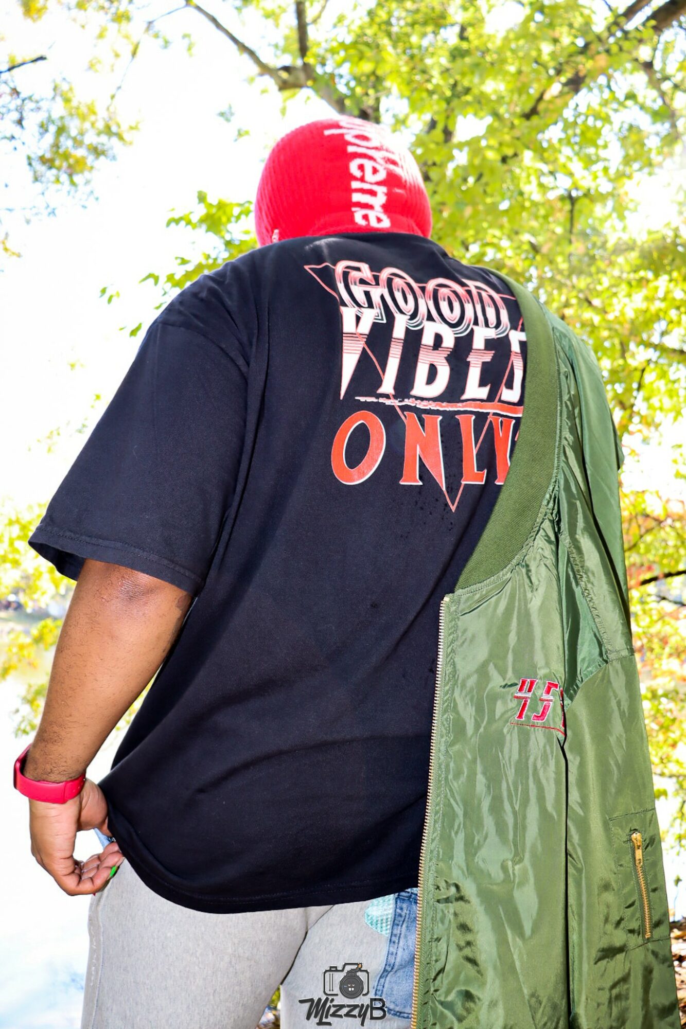 Person wearing a black T-shirt with colorful text, red beanie, and carrying a green bag outdoors with trees in background.