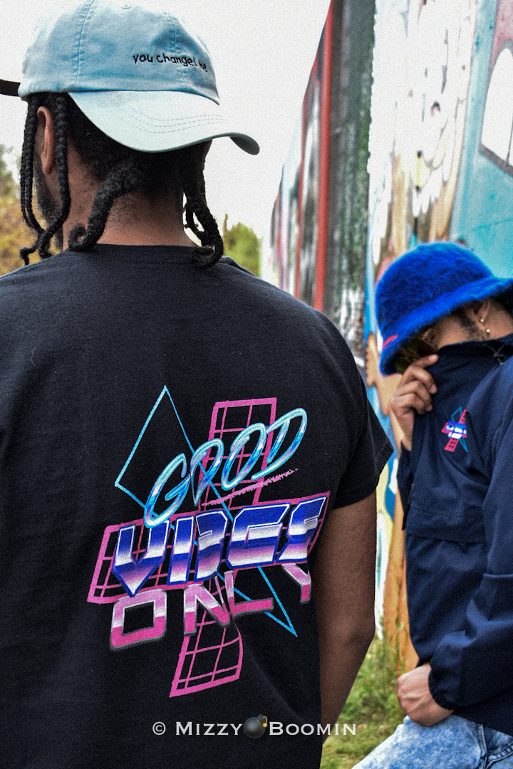 Two people with hats, one wearing a black T-shirt with colorful text, the other in a blue jacket and hat, outdoors.