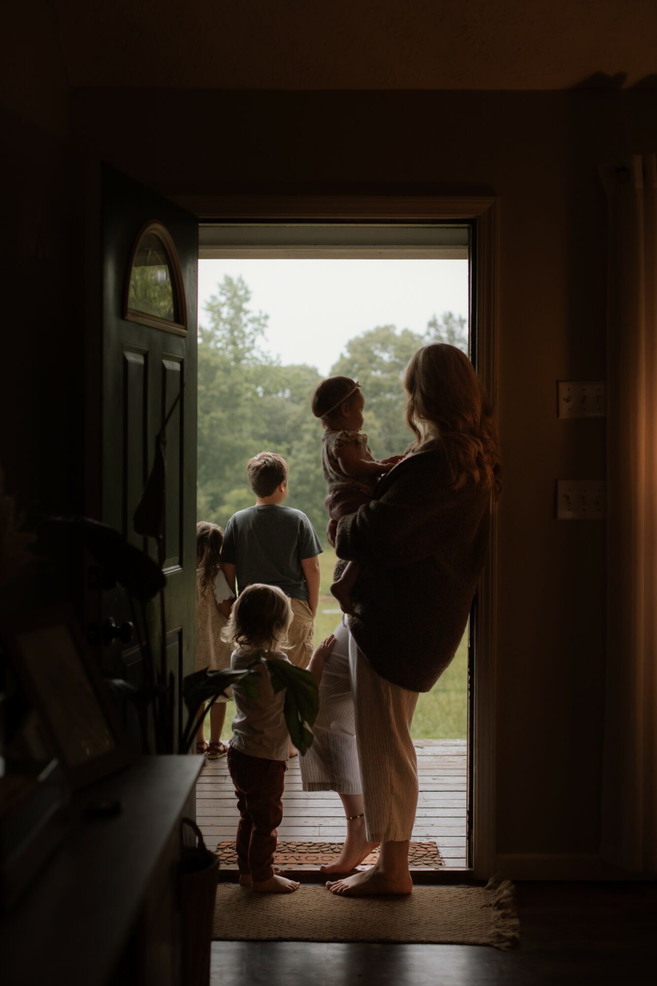 Person holding a child at doorway with other children outside, trees visible in background, interior dimly lit.