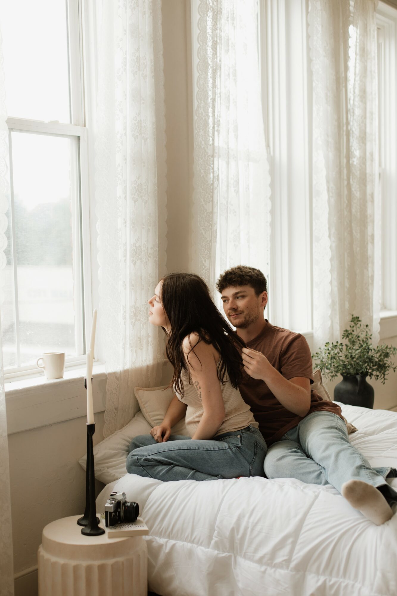 Two people sitting on a bed near a window with sheer curtains, with a lamp and camera on a side table.